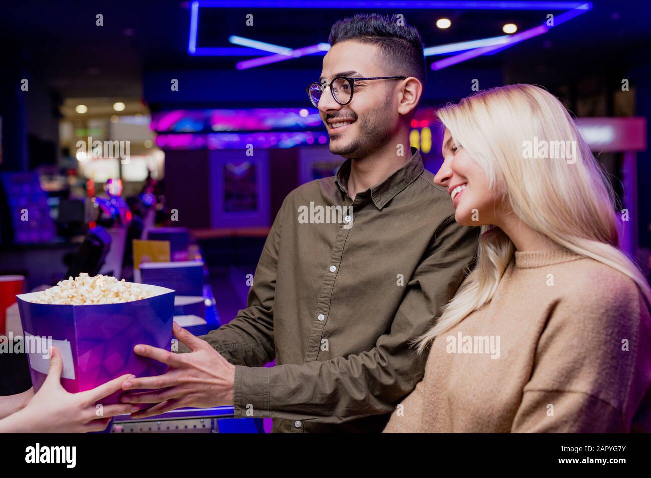 Jeune couple joyeux debout par comptoir de caissier tout en achetant du pop-corn au cinéma Banque D'Images