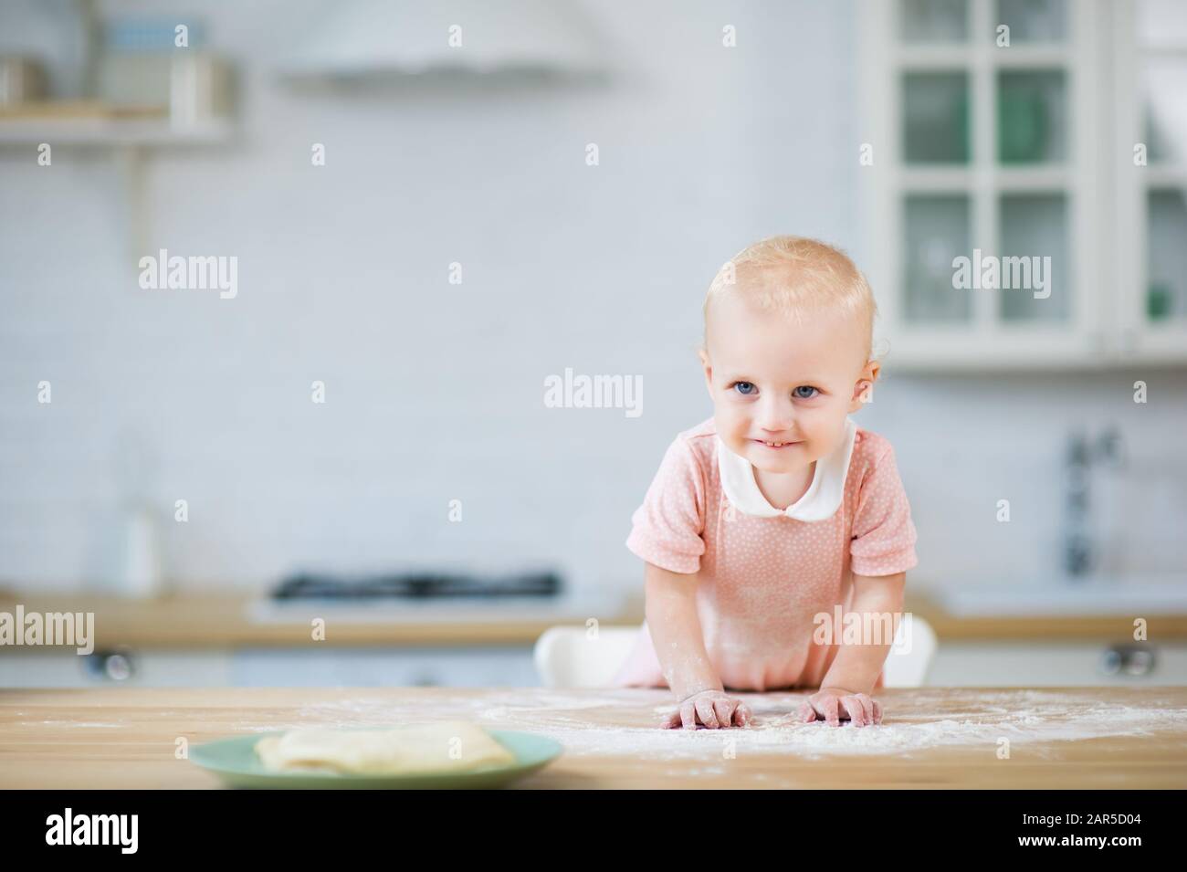 une fillette blonde aux yeux bleus sourit sly à l'appareil photo et se penche sur une table parsemée de farine Banque D'Images