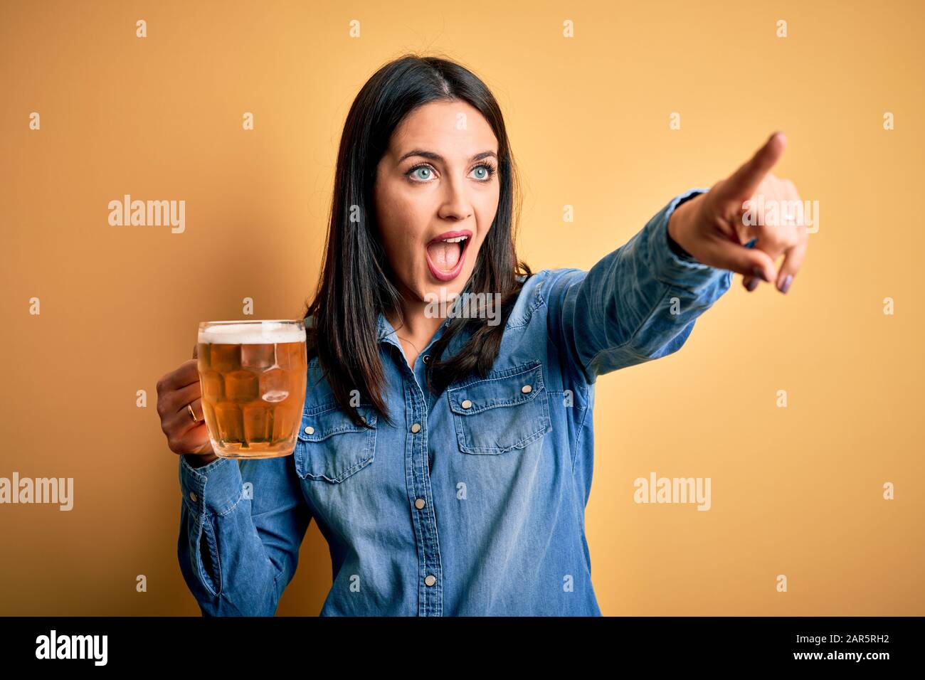 Jeune femme aux yeux bleus buvant du pot de bière debout sur fond jaune isolé Pointant avec le doigt surpris devant, bouche ouverte étonné expre Banque D'Images