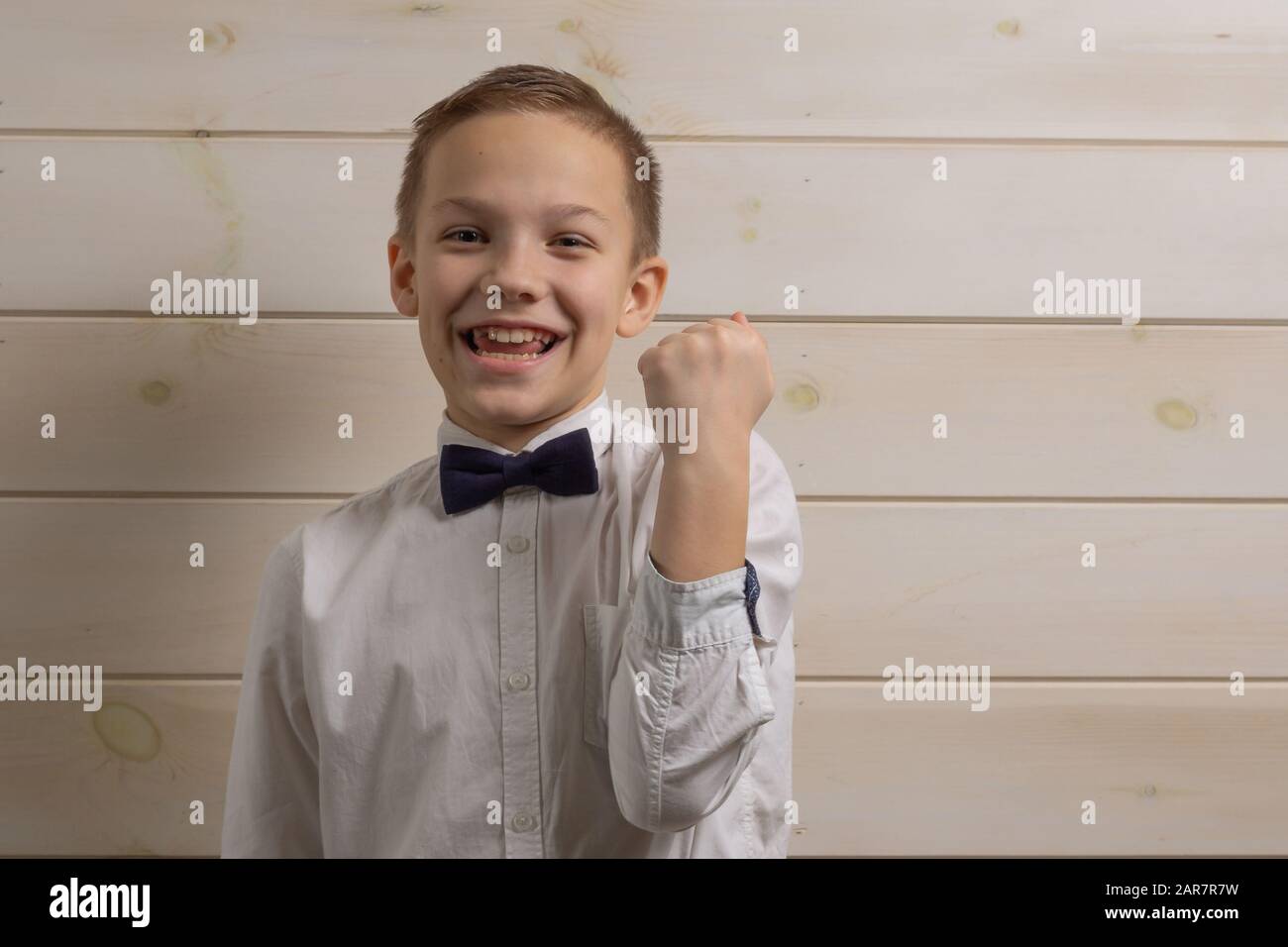Un garçon de 10 ans aux cheveux justes dans une chemise blanche avec noeud  papillon à nouer souriait sur le fond d'un mur en bois. Geste oui,  victoire, je l'ai fait Photo