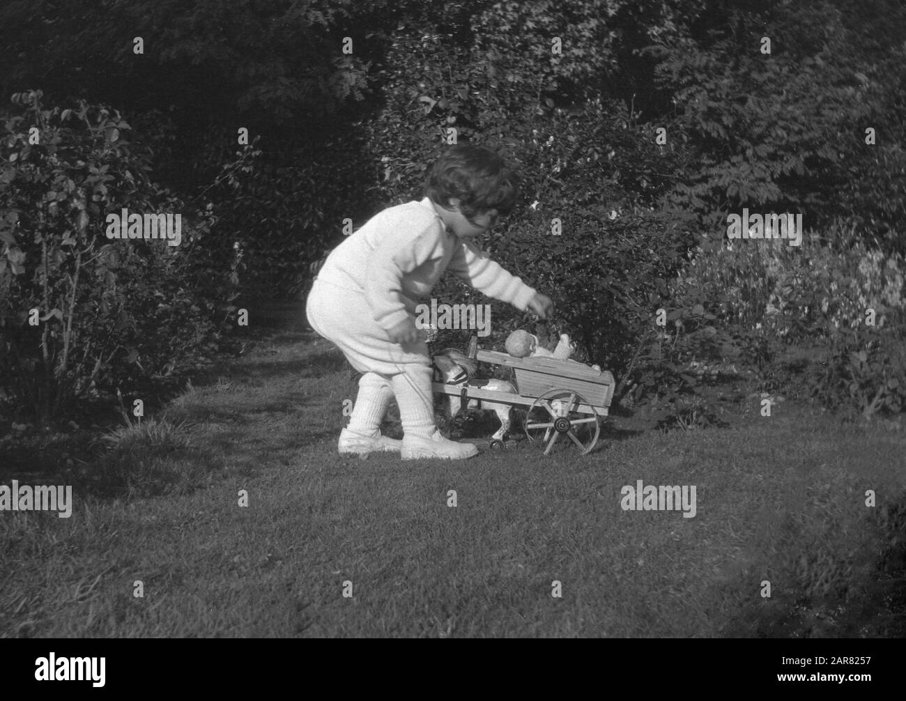 Années 1920, historique, une petite fille dehors dans un jardin jouant avec sa poupée assise dans un cheval en bois jouet & charrette, Angleterre, Royaume-Uni. Banque D'Images