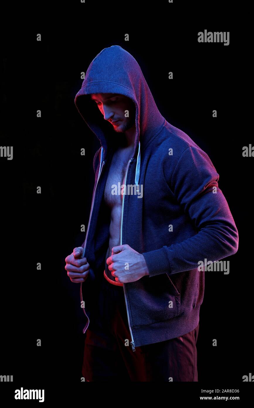 Jeune athlète masculin dans un sweat à capuche bleu foncé non zippé debout dans l'obscurité Banque D'Images