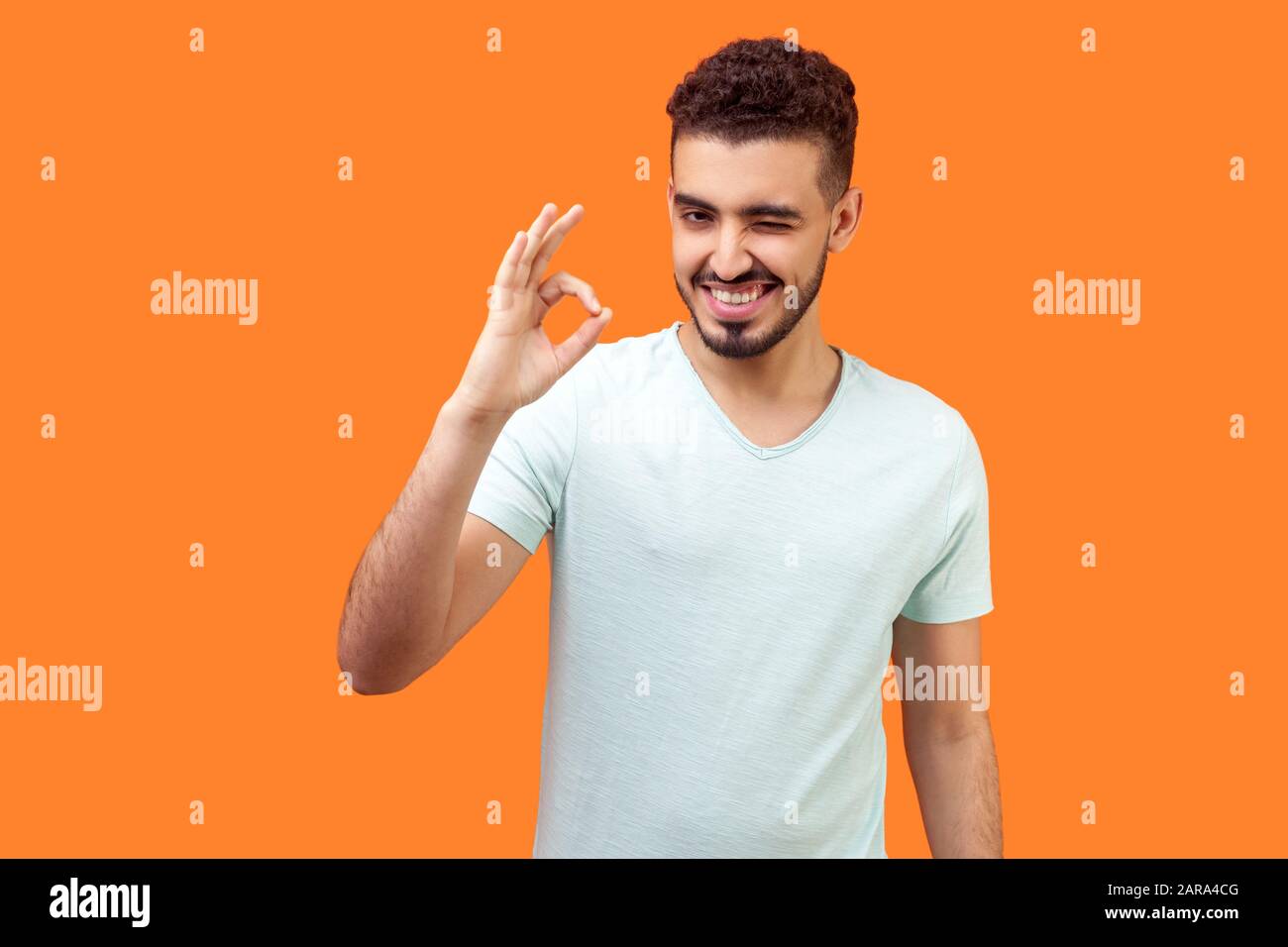 Je vais bien ! Portrait d'un homme espiègle et optimiste avec une barbe dans un t-shirt blanc montrant un geste correct du doigt et un clin d'œil à l'appareil photo, flirtant. Banque D'Images