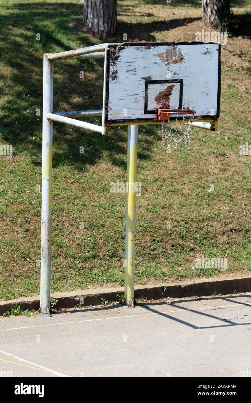 Un vieux panier de basket-ball délabré avec un panneau en bois cassé  fissuré et un cadre en métal fort entouré d'un terrain de basket-ball pavé  et d'herbe fraîchement coupée Photo Stock -