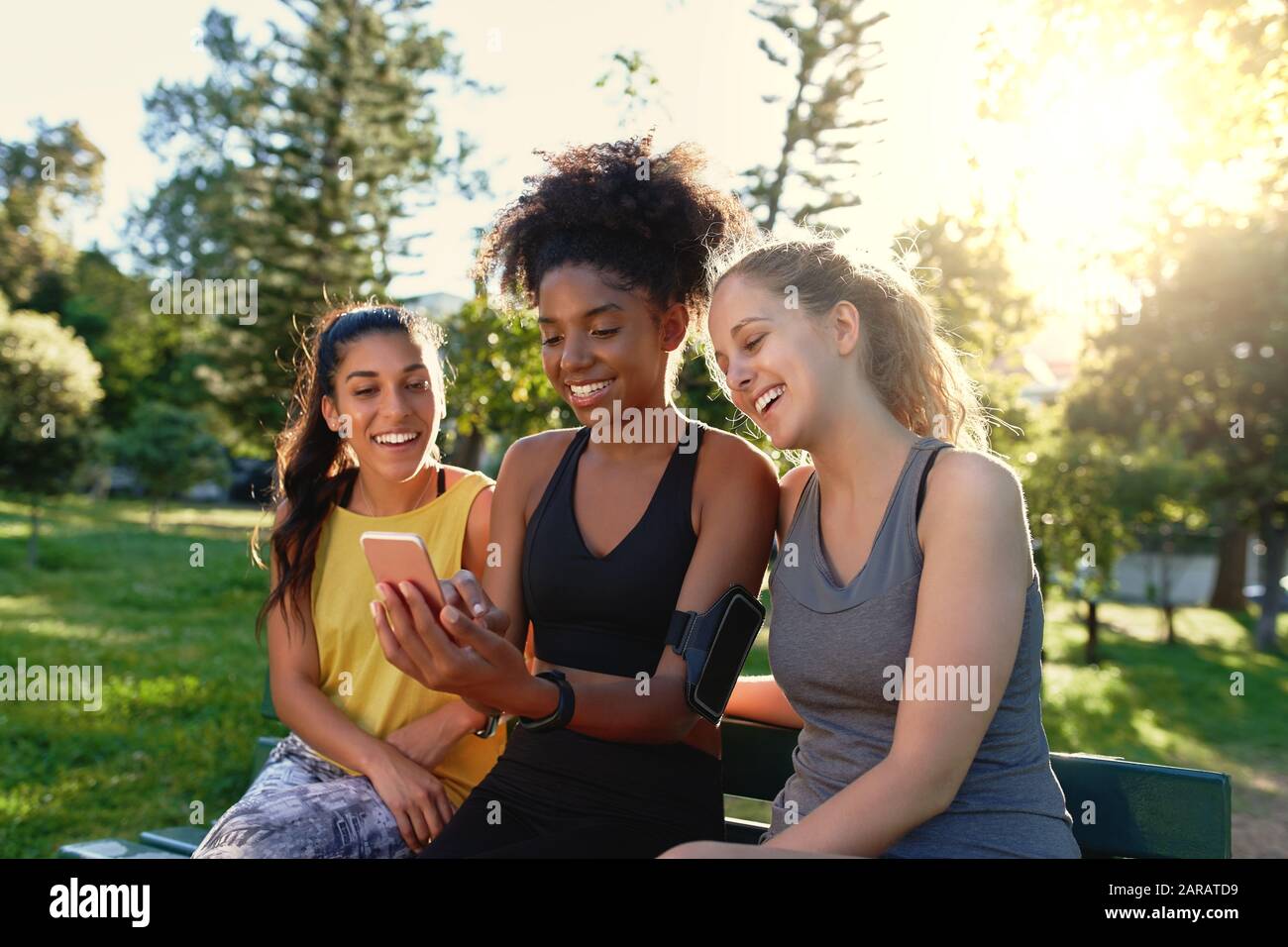 Des jeunes amis sportifs et variés assis ensemble sur un banc en regardant le téléphone mobile dans le parc le matin - heureux amis de forme physique souriant et Banque D'Images