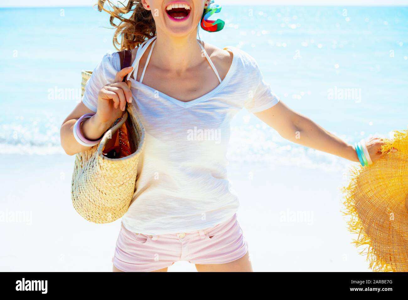 Gros plan sur une heureuse femme moderne dans un t-shirt blanc avec sac de paille de plage et grand chapeau sautant sur la rive de l'océan. Banque D'Images