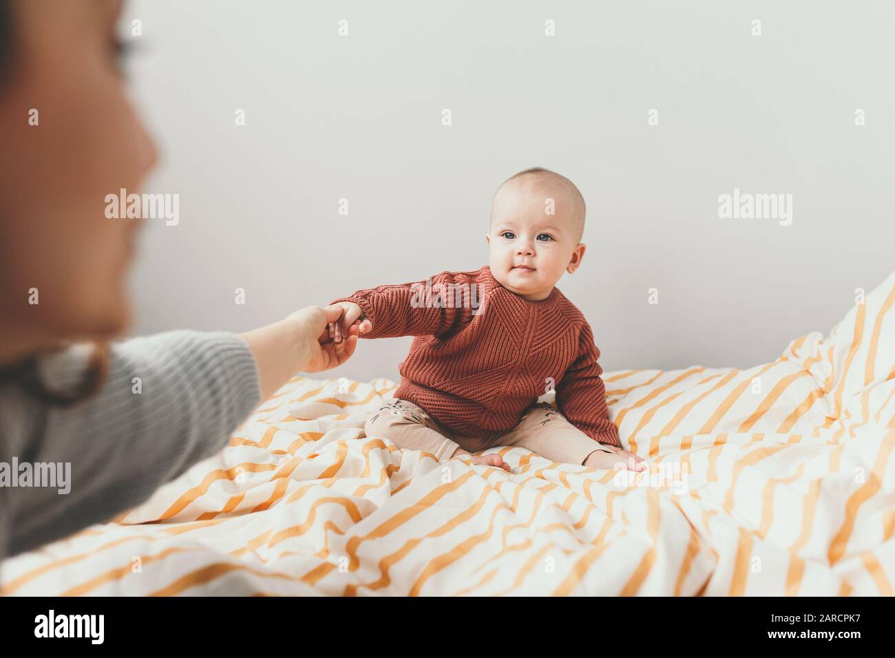 Belle petite fille de bébé sur le lit dans un confortable pull brun souriant. Concept de maternité et d'enfance. Adorable petite fille de six mois posée sur le mauvais et regardant dans l'appareil photo. La mère tient une petite main. Banque D'Images