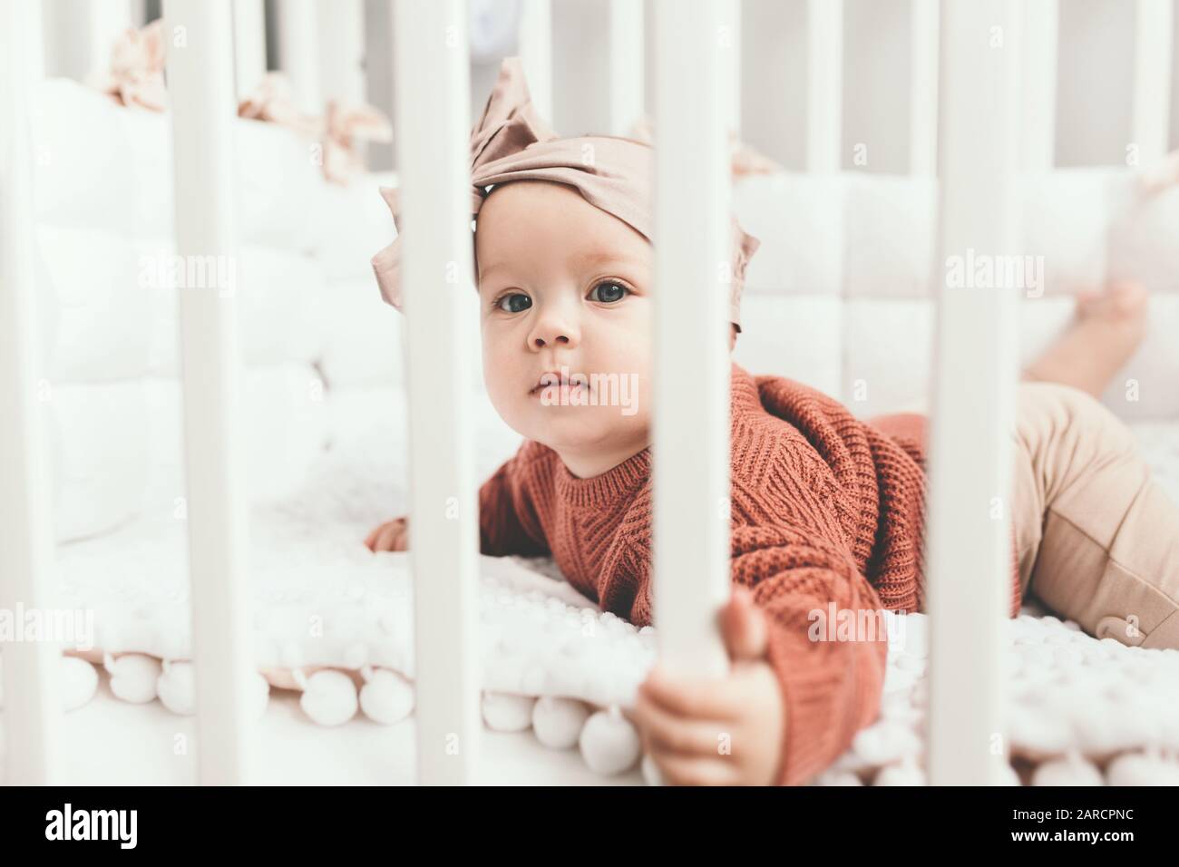 Belle petite fille dans un lit de bébé dans une salle de lumière confortable dans un pull de tons à la mode. Apprend à se tenir au soutien. Petite fille gaie debout dans le lit de bébé à la maison Banque D'Images