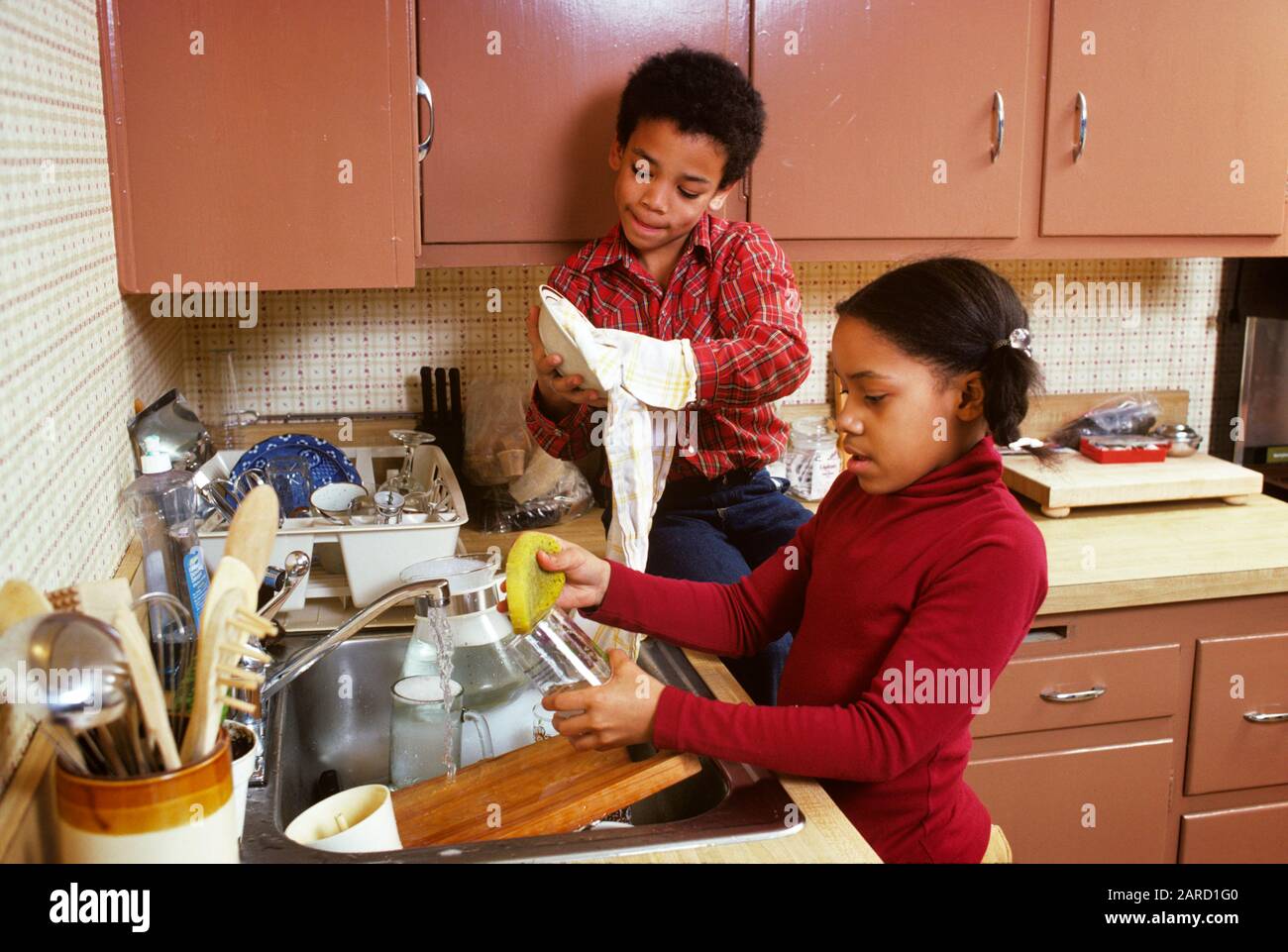 ANNÉES 1970 1980 PETIT FRÈRE ET SŒUR AFRO-AMÉRICAIN ET FILLE ENSEMBLE VAISSELLE DE LAVAGE PAR PETIT GARÇON ASSIS SUR LE COMPTOIR DE CUISINE - KJ8830 PHT001 LES FRÈRES FILLES HARS ACCUEIL VIE COPIE ESPACE AMITIÉ DEMI-LONGUEUR HOMMES FRÈRES SŒURS HAUTES SOEURS AFRO-AMÉRICAINS ET ETHNICITÉ NOIRE HAUT LIEN FRÈRE SOUTIEN COOPÉRATION JEUNES PRÉ-ADOLESCENCE PRÉ-ADOLESCENCE L'ÂGE DES FILLES A DÉMODÉ LES AFRO-AMÉRICAINS Banque D'Images