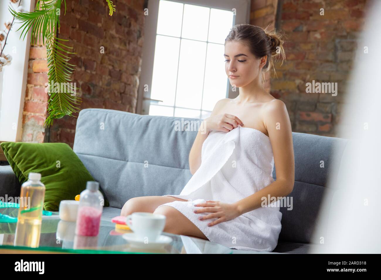 Journée de beauté pour vous-même. Une femme aux cheveux longs porte une serviette faisant sa routine quotidienne de soin de la peau à la maison. Repose sur un canapé, pour préparer l'hydratant, masser. Concept de beauté, de soins de soi, de cosmétiques. Banque D'Images