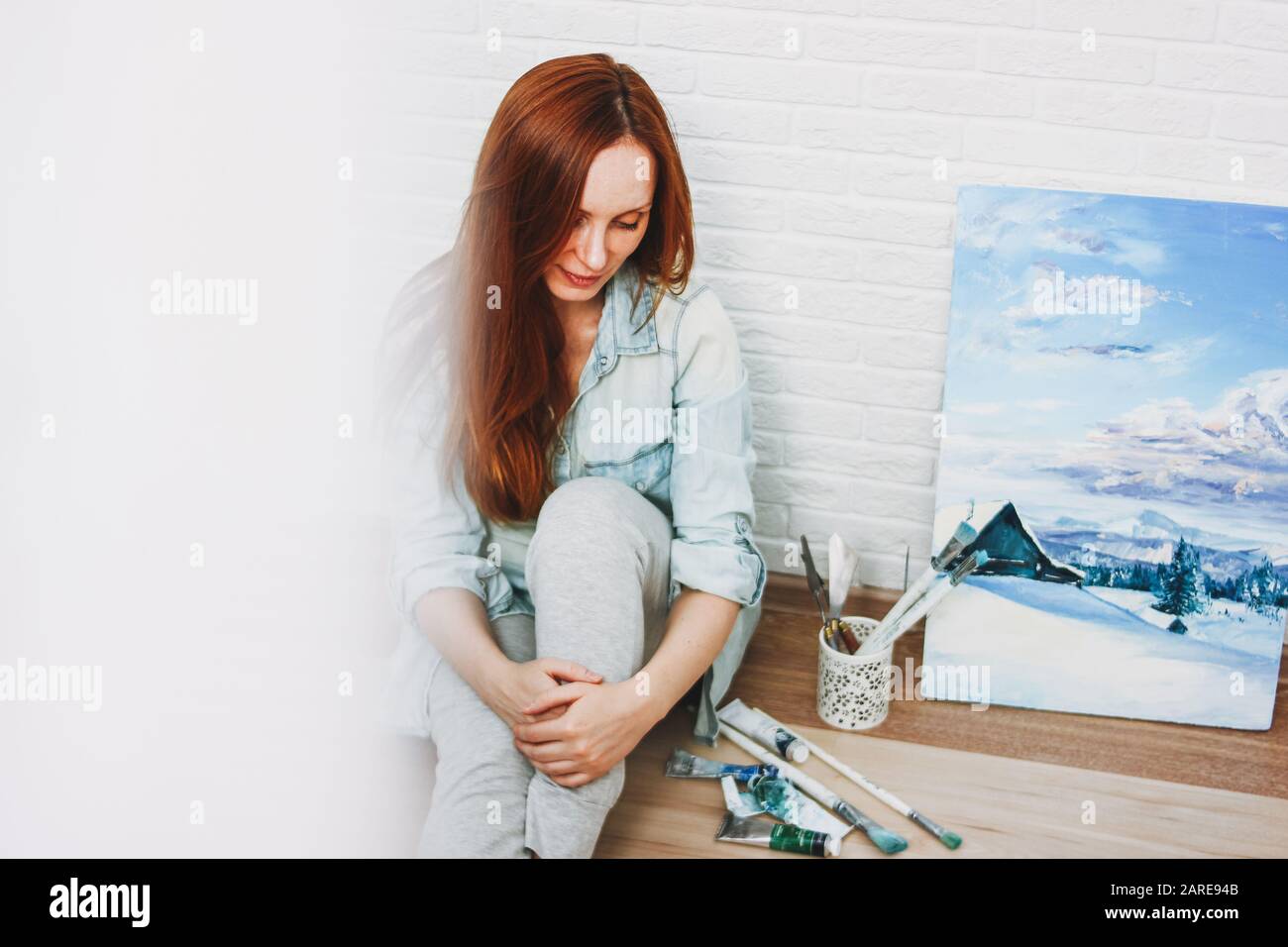 Jeune femme artiste avec de longs cheveux rouges assis près de son travail d'art photo au sol au studio Banque D'Images