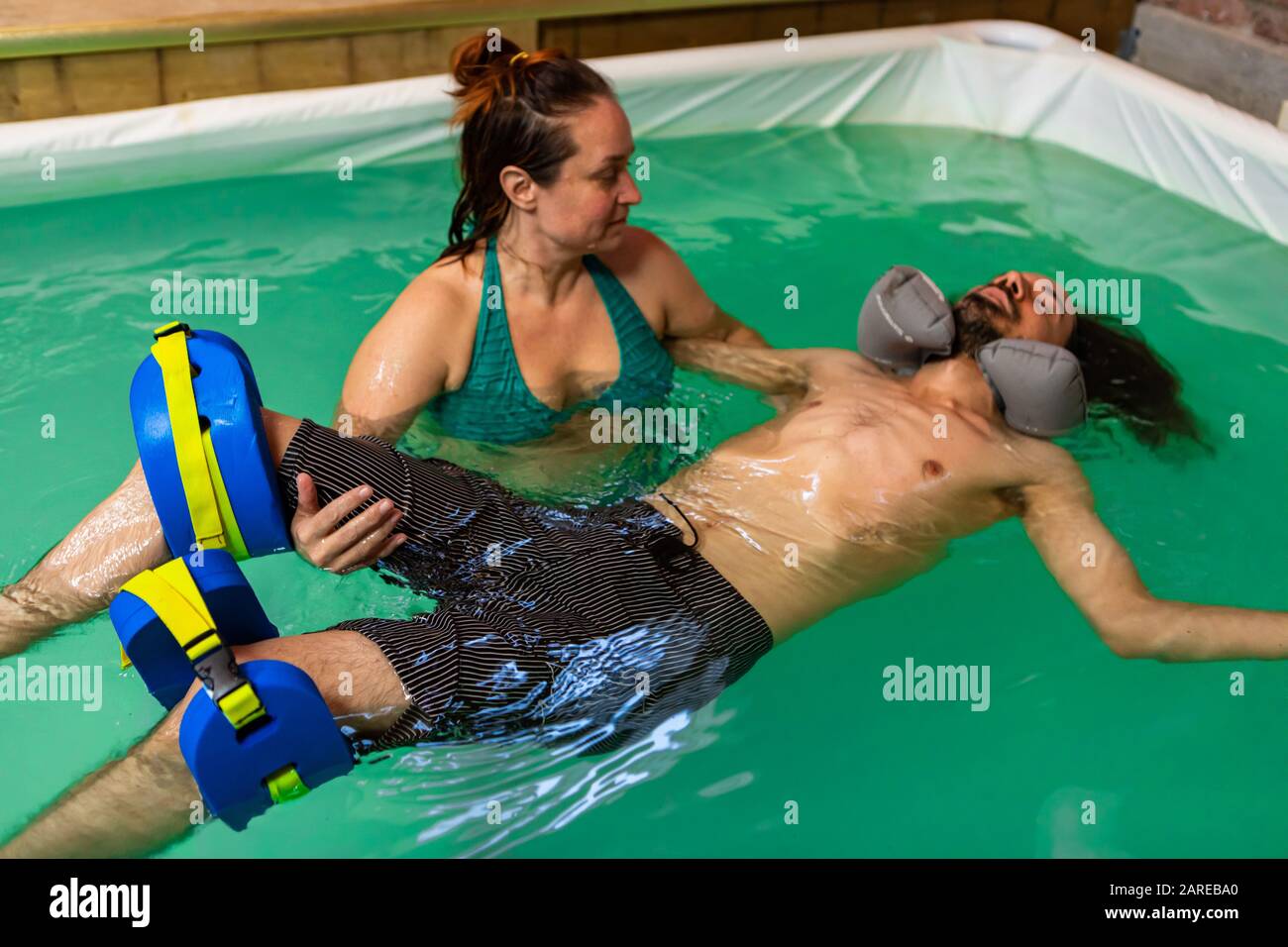 Jeune homme allongé sur l'eau dans la piscine avec flotteur de cou et soutien de jambe obtenir le traitement d'aqua pour un avec un entraîneur expérimenté de femme Banque D'Images