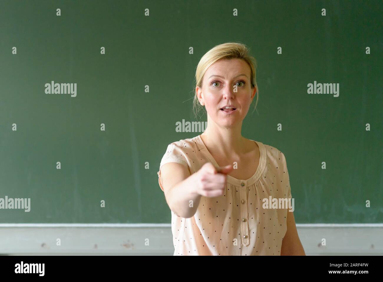 Professeur d'école debout devant un tableau noir dans la salle de classe pointant avec son doigt vous disant Banque D'Images