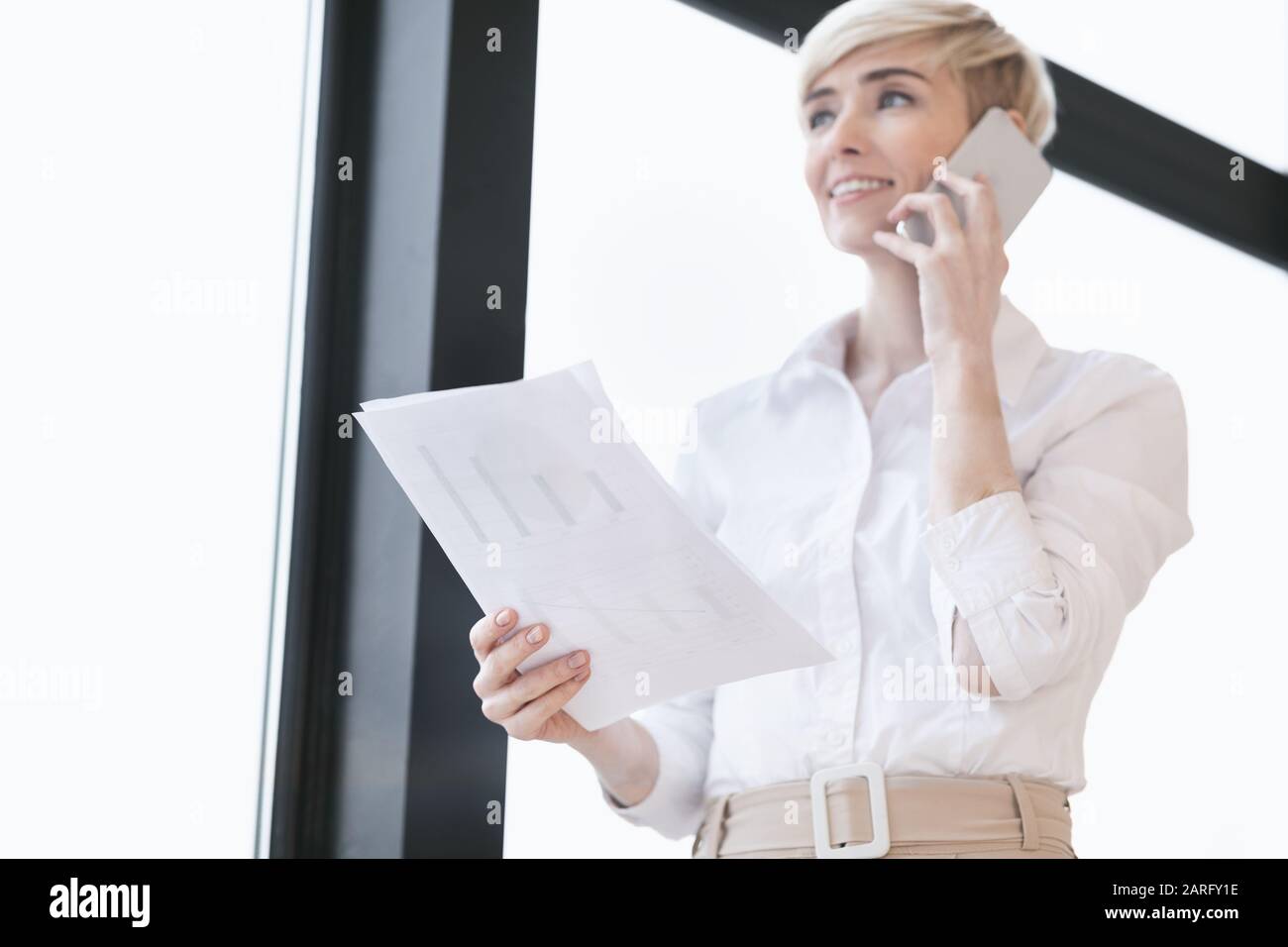 Femme D'Affaires Parlant Sur Cellphone Debout Près De La Fenêtre Dans Le Bureau Moderne Banque D'Images