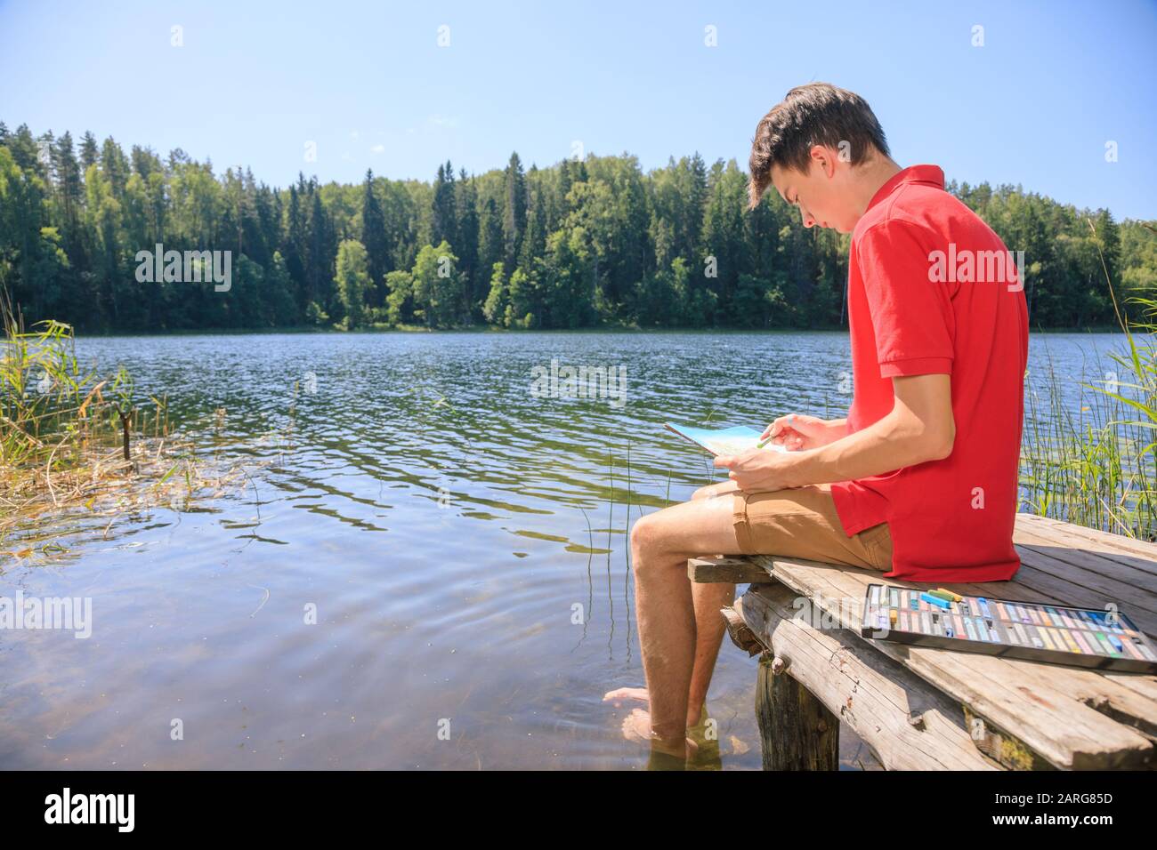 Jeune garçon assis sur une jetée en bois au bord d'un lac forestier avec des crayons pastel - plein air concept de scotching Banque D'Images