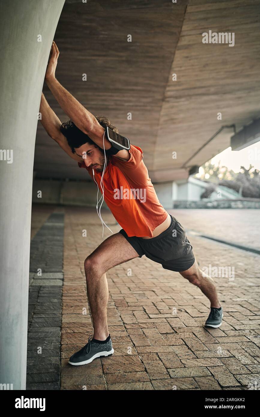 Beau jeune homme dans des vêtements de sport avec écouteurs et brassard se réchauffant en étirant son bras et sa jambe pendant la course du matin Banque D'Images
