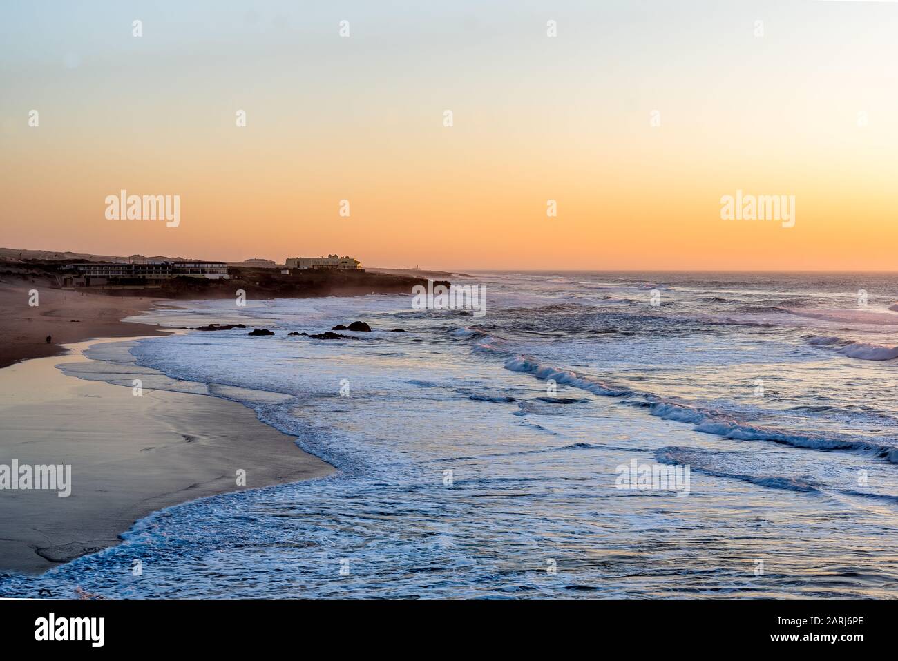 Les vagues de l'océan Atlantique qui s'étendent sur la plage Guincho - Praia do Guincho - sur la côte de Sintra au Portugal au coucher du soleil avec des bâtiments sur le promontoire en gro Banque D'Images