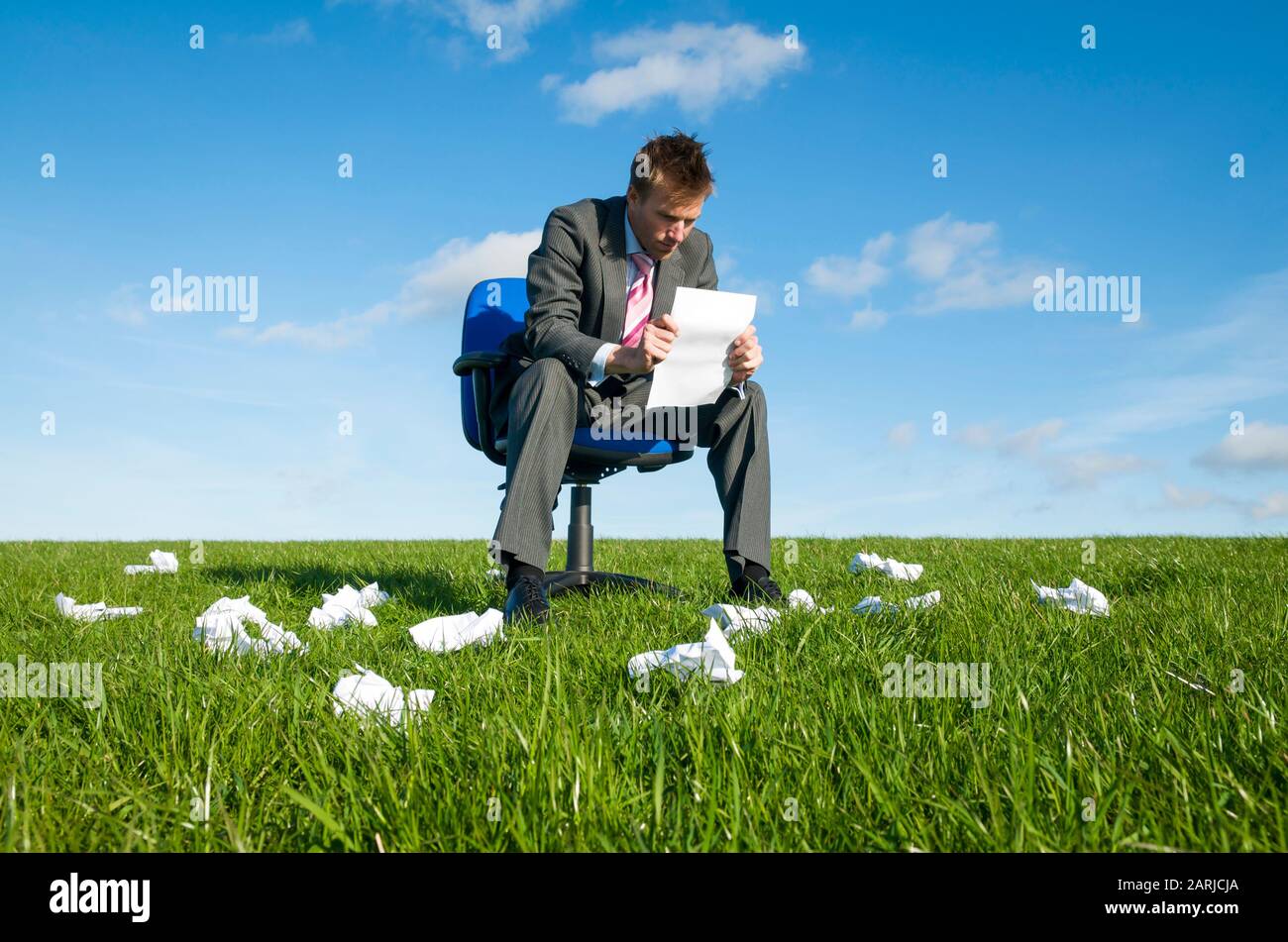 Homme d'affaires lisant une feuille de papier sur une chaise de bureau, froissé des boules de courants d'air précédents dispersés dans la prairie autour de lui Banque D'Images