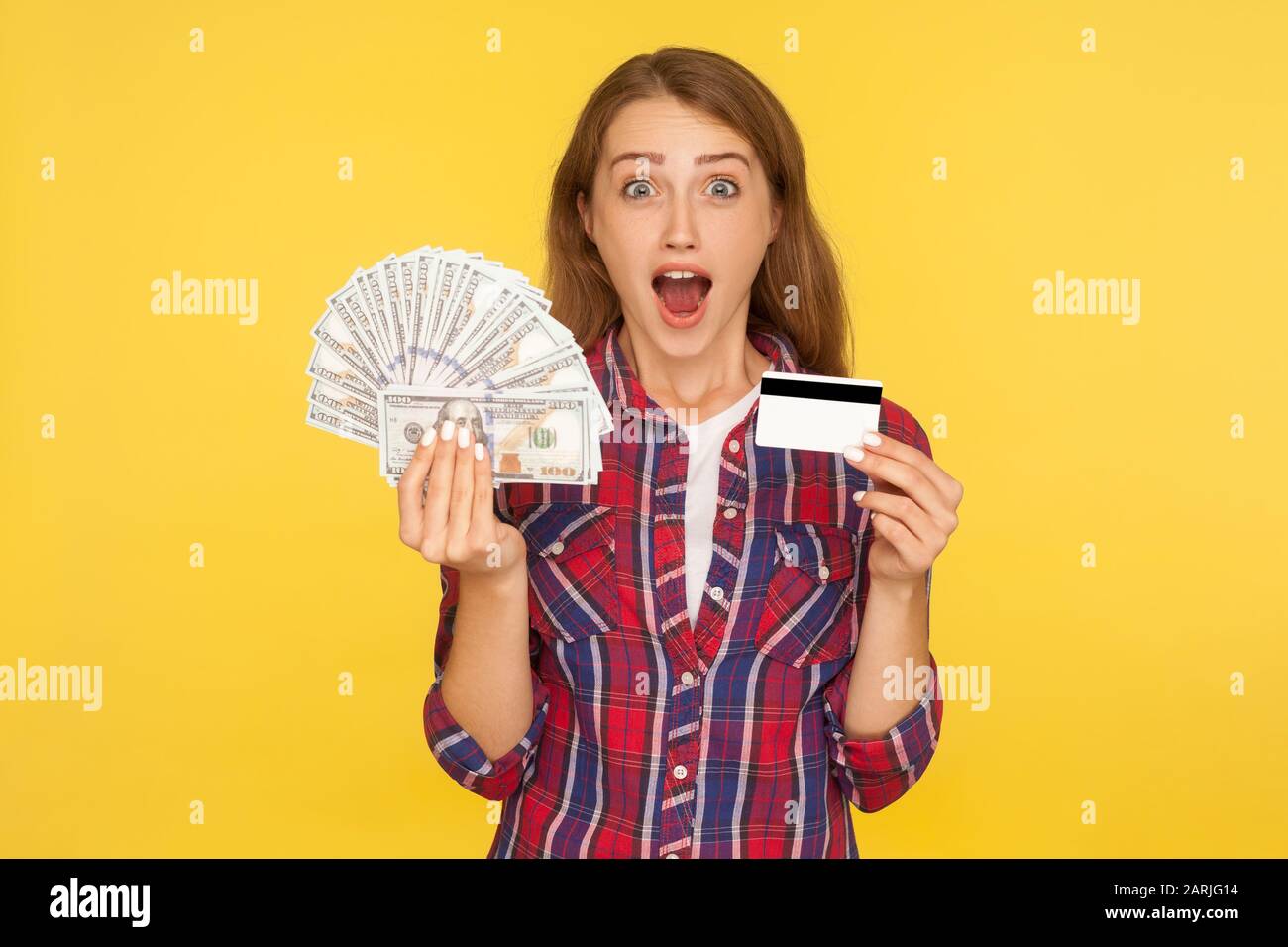 Une heureuse fille de gingembre enchantée dans une chemise debout avec une carte bancaire en plastique et des billets en dollars dans ses mains, regardant surpris par l'appareil photo. Indo Banque D'Images