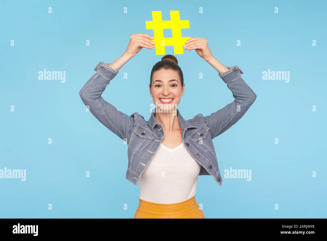 Jolie fille joyeuse avec un petit pain dans un ensemble élégant tenant le grand symbole hashtag jaune sur la tête et souriant à l'appareil photo, signe de hachage des médias sociaux, bloggi Banque D'Images