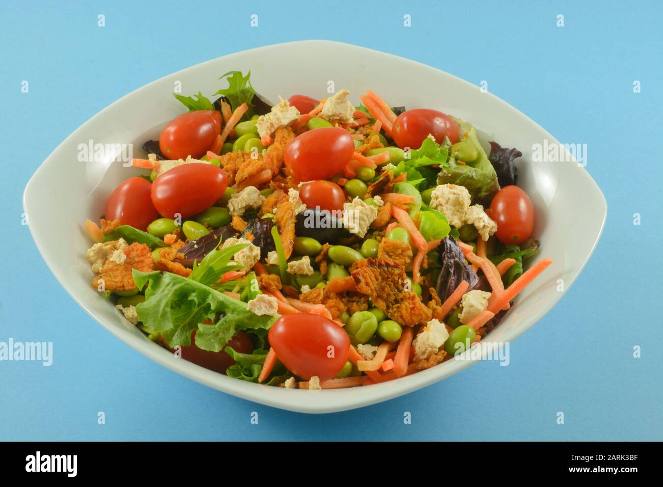 Salade saine avec des légumes frais mélangés, des tomates de raisin, des carottes de matchstick, de l'edamame, du fromage et des poivrons rouges croustillants dans un bol blanc sur fond bleu Banque D'Images