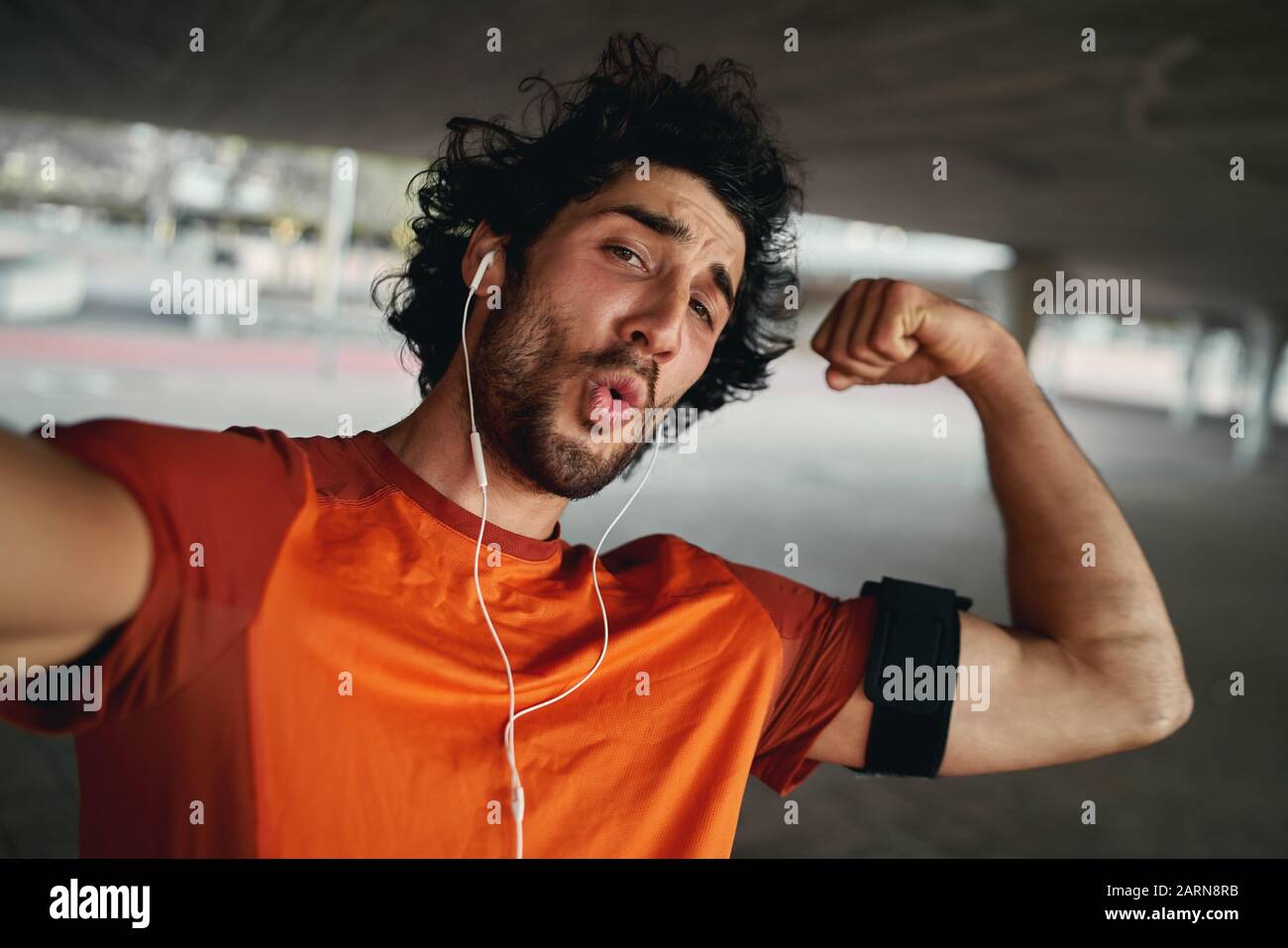 Un jeune sportif à la coupe attrayante avec écouteurs qui fait du visage drôle fléchissement de ses muscles tout en prenant un selfie dehors Banque D'Images