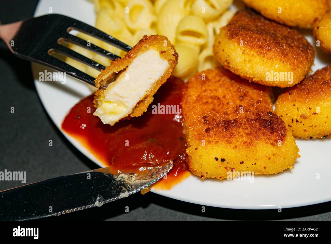 Un morceau de pépites sur une fourchette et une plaque blanche avec des pépites de poulet frit, des pâtes et du ketchup sur un fond sombre. Gros plan Banque D'Images