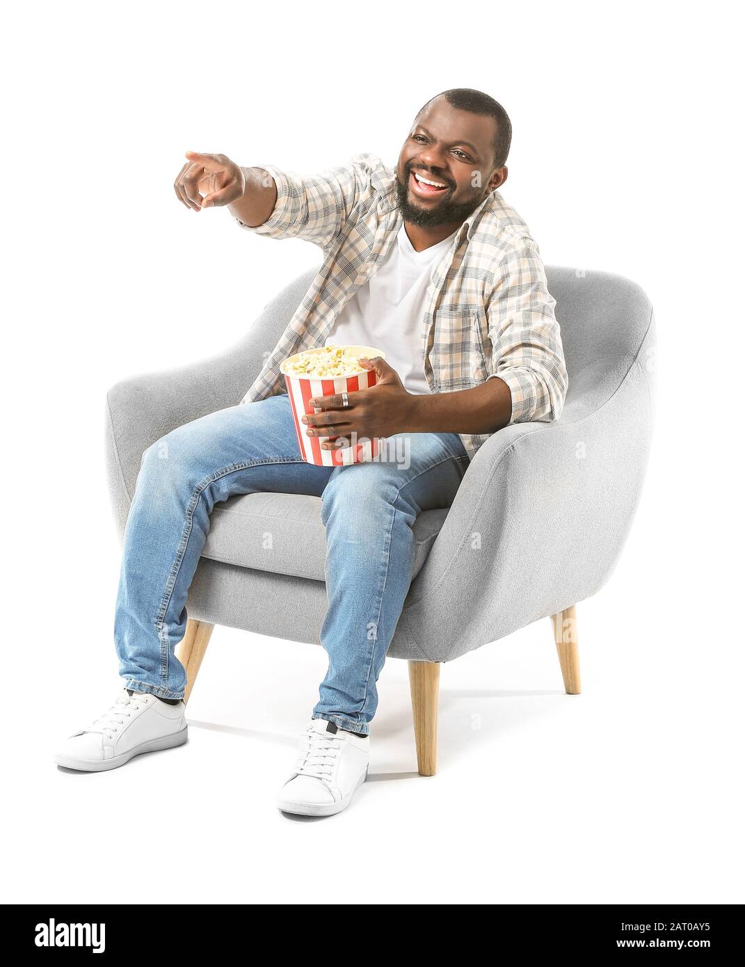 Homme afro-américain avec pop-corn regardant la télévision tout en étant assis dans un fauteuil sur fond blanc Banque D'Images