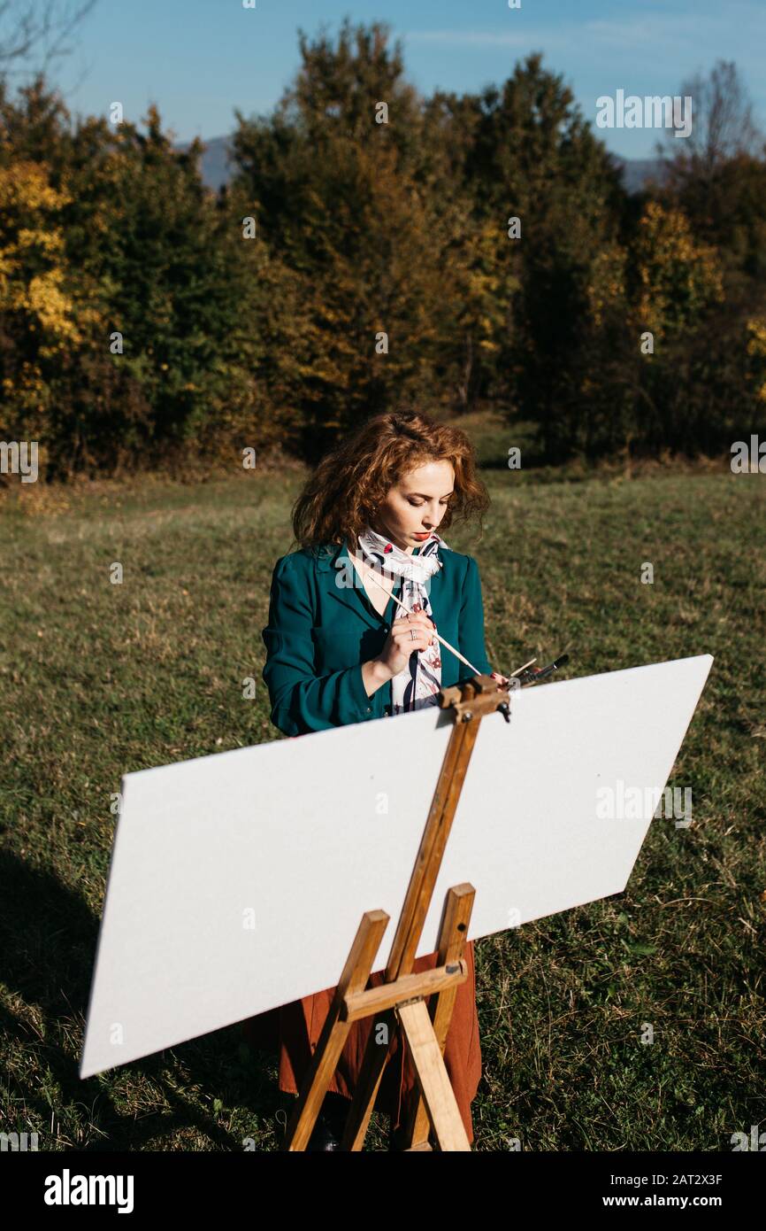 sortez l'artiste féminine qui travaille sur la peinture à l'extérieur, dans le paysage d'automne. Elle est devant la toile et le dessin.Elle tient des peintures à l'huile, brosse d'artiste Banque D'Images