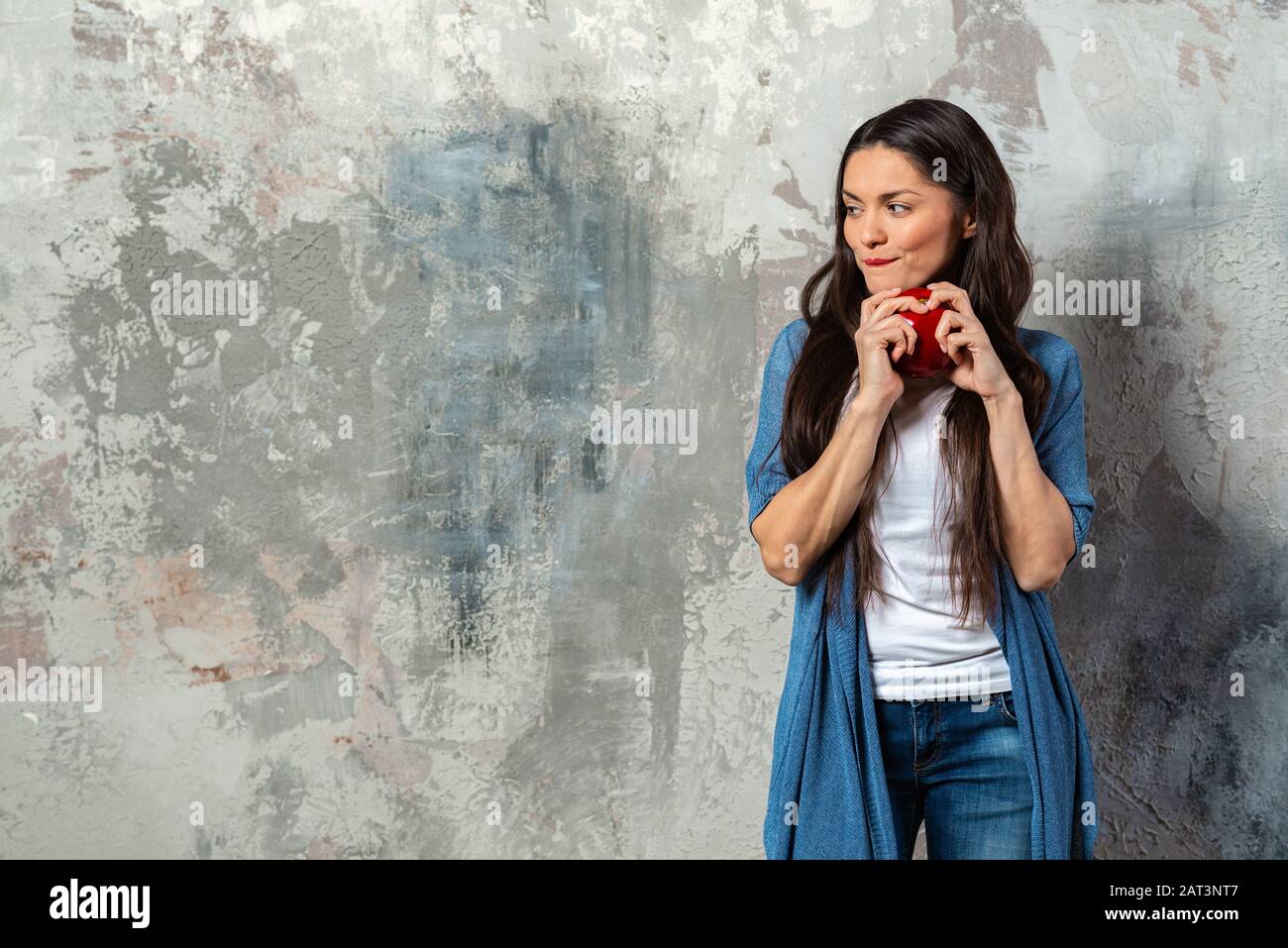 Un portrait d'une demi-longueur de femme tenant une grosse pomme rouge à la fois bras et regardant loin joueusement. Se tient sur l'arrière-plan abstrait Banque D'Images