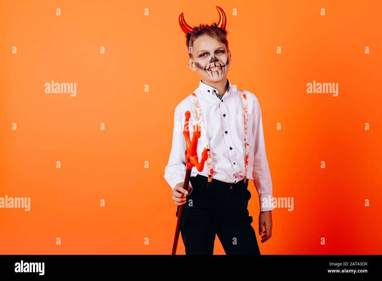 Diable garçon debout demi-tour contre un fond orange dans le maquillage de mascarade. Concept d'Halloween Banque D'Images