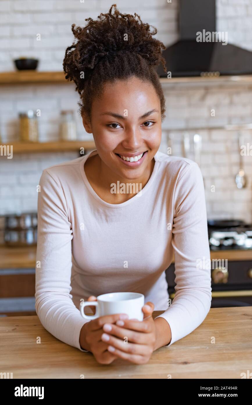 Jolie fille mulatto se trouve à la table avec une tasse de thé ou de café du matin et sourire regardant l'appareil photo Banque D'Images