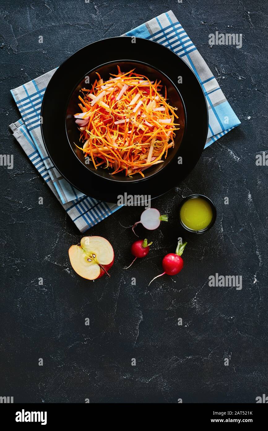 Salade fraîche de radis et de pomme aux carottes saupoudrée de graines de quinoa et de zeste de citron dans un bol noir sur une table en bois avec ingrédients, vue verticale Banque D'Images