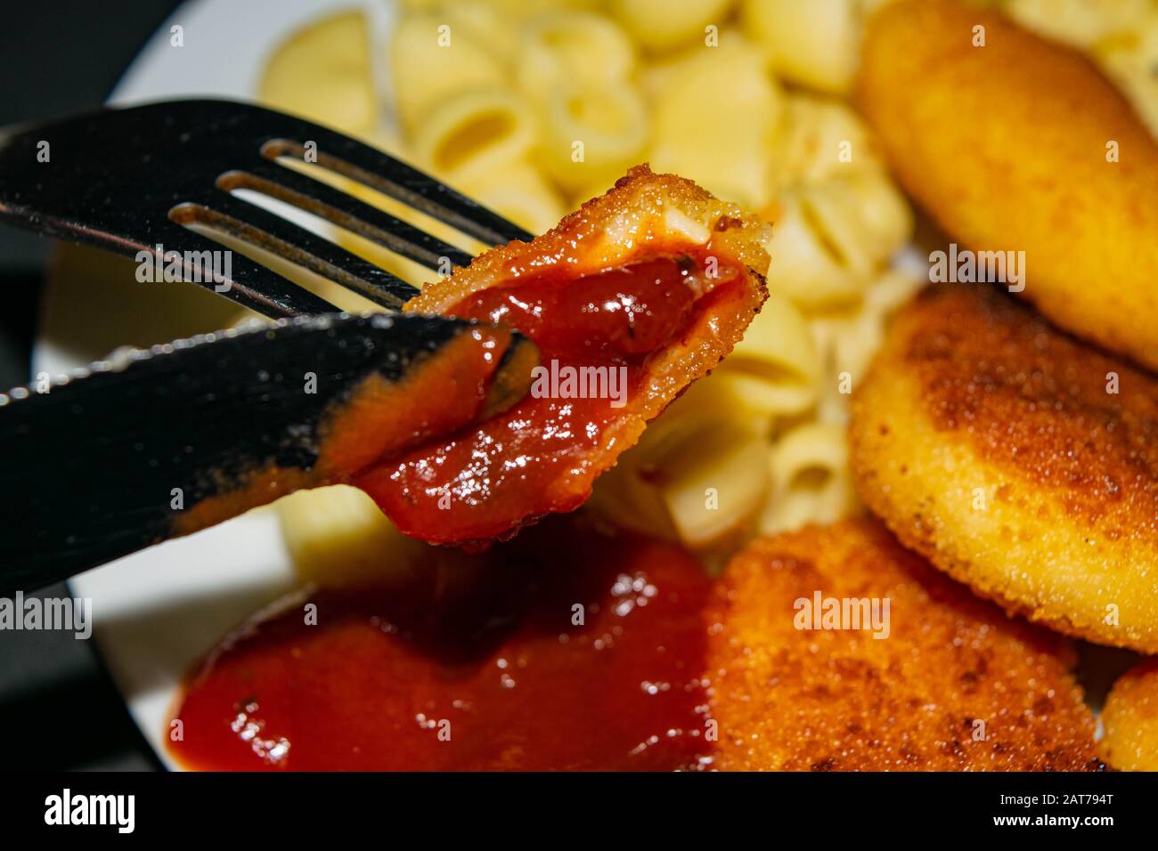 Un morceau de pépites avec ketchup sur une fourchette et une plaque blanche avec des pépites de poulet frits, des pâtes et du ketchup sur un fond sombre. Gros plan Banque D'Images