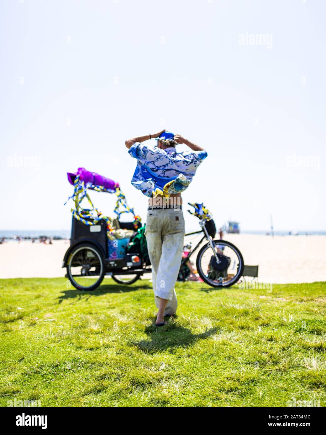 Homme Solitaire Dans Des Vêtements Aux Couleurs Vives Et Festives Marchant Sur Grass Vers Vélo Décoré Sur Venice Beach Surplombant L'Océan Pacifique. Banque D'Images