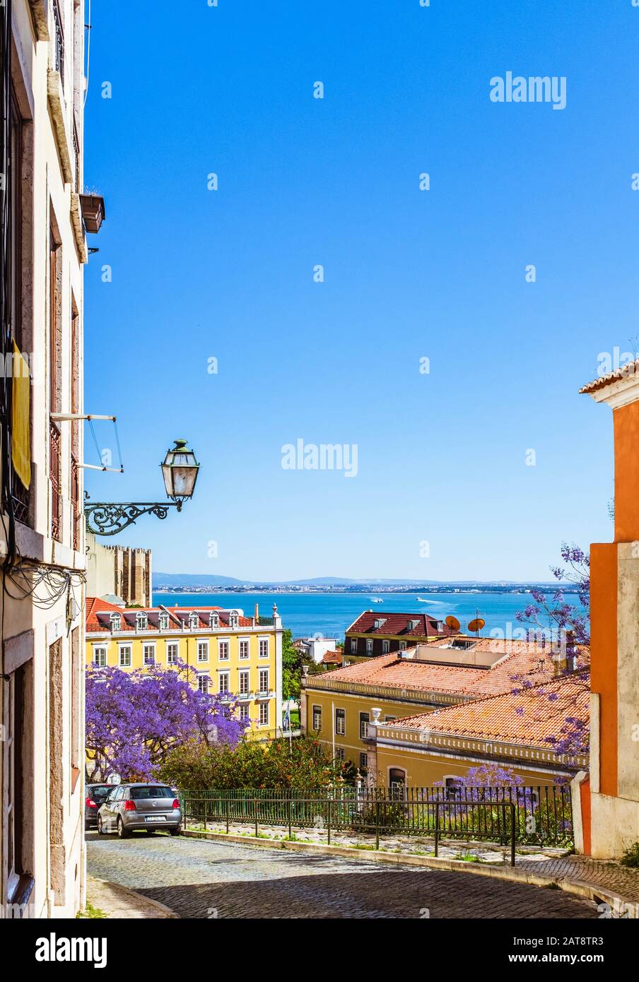 Une rue pavée abrupte dans le quartier d'Alfama lors d'une journée de printemps ensoleillée avec le Tage sur fond. Banque D'Images