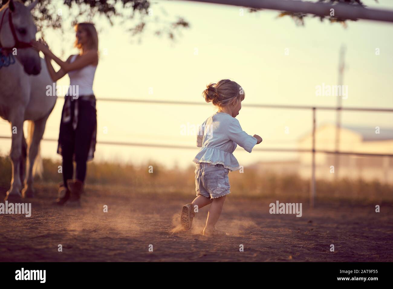 Petite fille qui court sur le ranch, alors que la maman est en train de petter un cheval. Divertissement sur la campagne, coucher de soleil heure d'or. Concept de la nature de la liberté. Banque D'Images