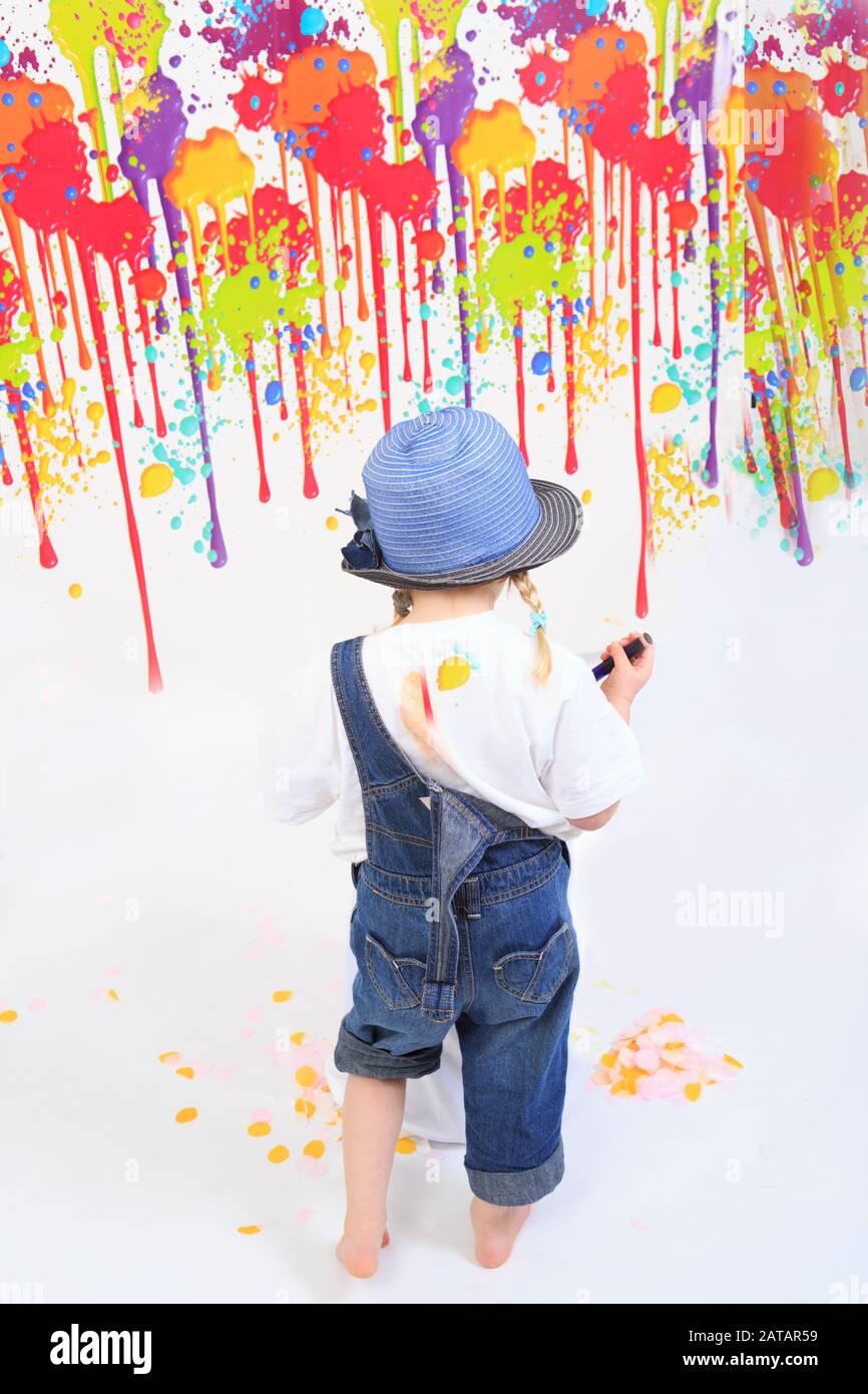 Peinture pour jeunes enfants, vue arrière d'une fille portant des salopettes regardant ses taches de peinture sur un mur blanc. Banque D'Images