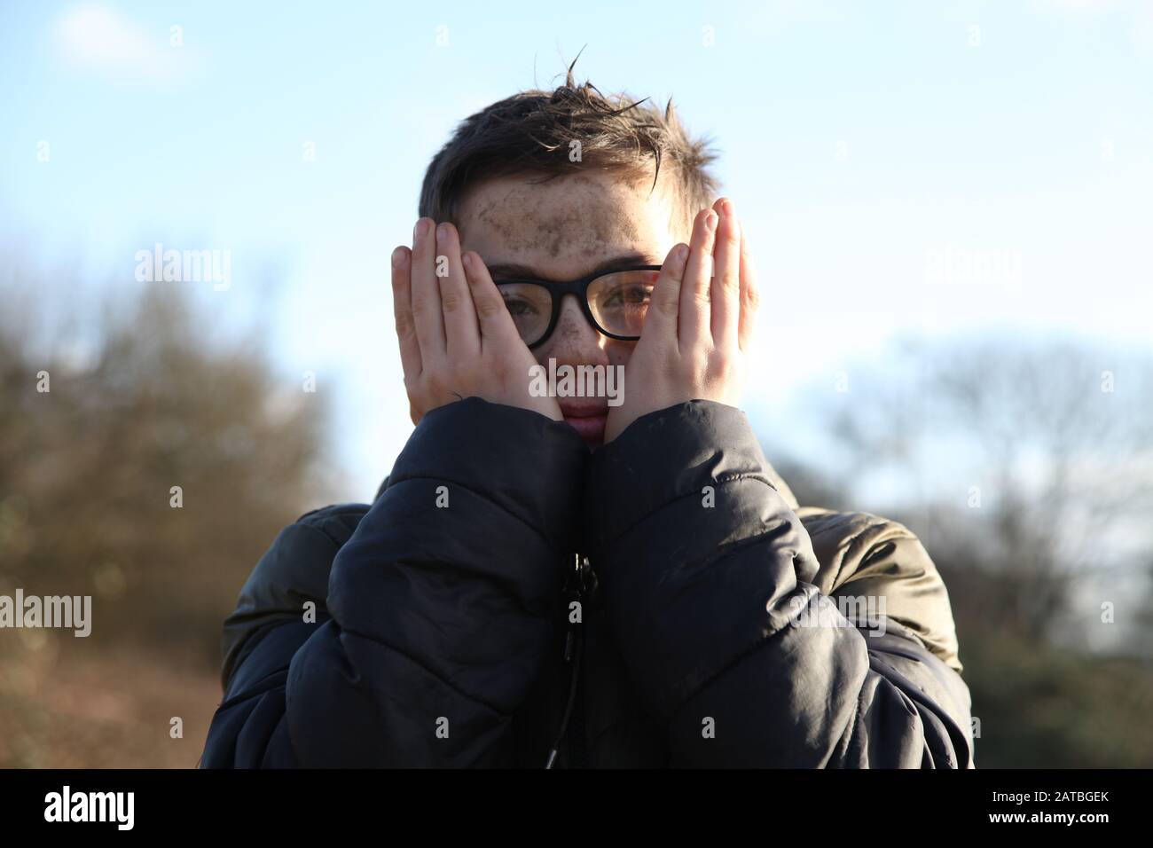 Un jeune garçon portant des lunettes de 12 ans couvre son visage boueux avec ses mains, face à l'appareil photo Banque D'Images