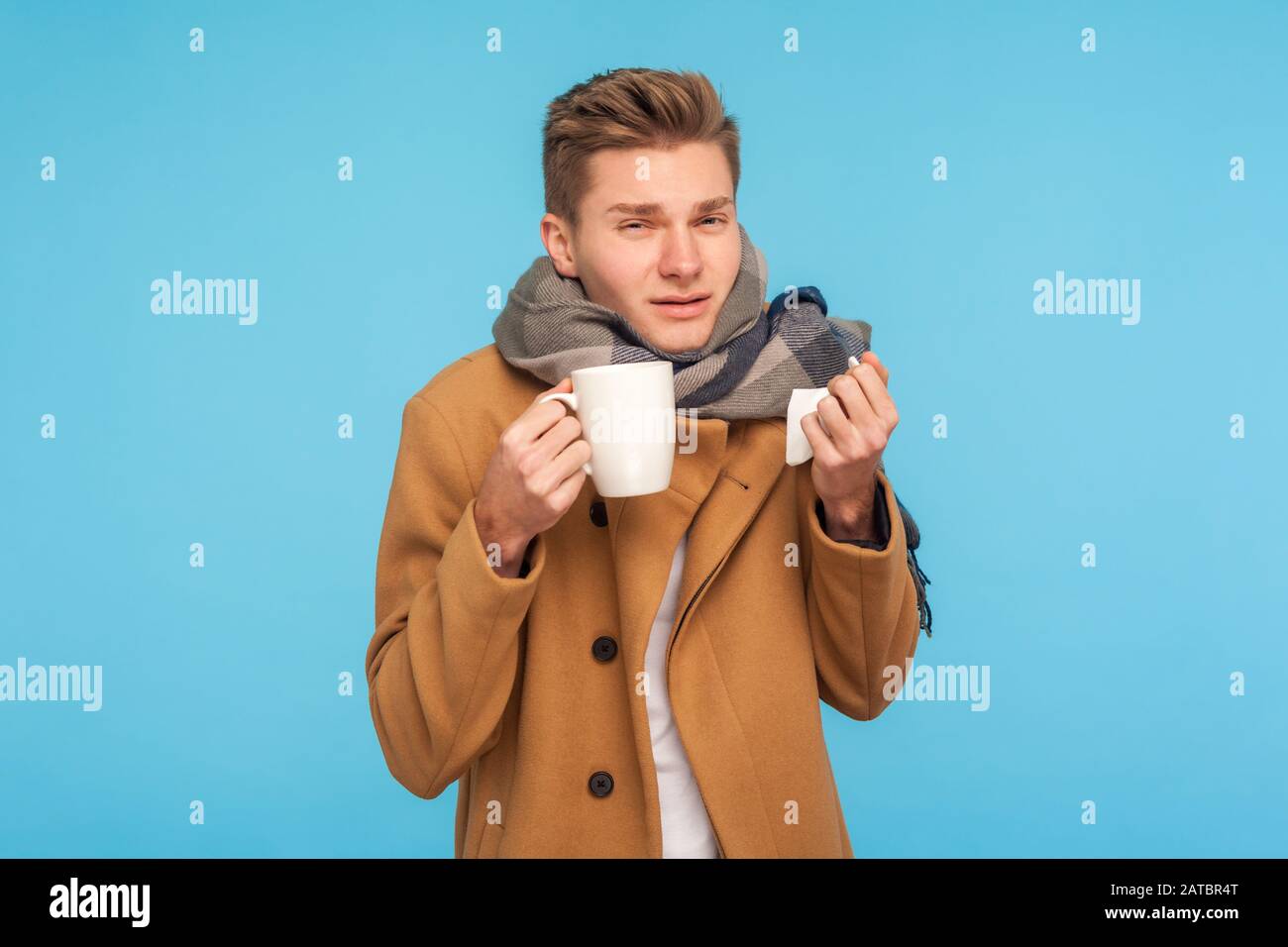 Traitement à domicile. Homme malade de la grippe en automne manteau et écharpe tenant mug et tissu pour le nez liquide, prenant des médicaments avec thé chaud, souffrant de fièvre, maux de tête. Banque D'Images