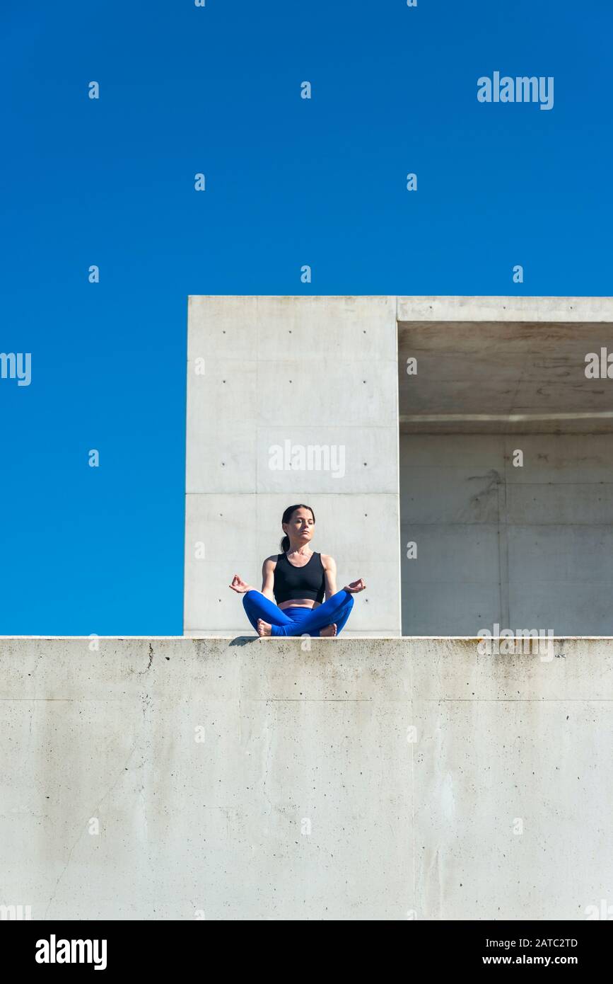 femme pratiquant le yoga et la méditation assis sur un mur à l'extérieur avec un fond d'architecture moderne. Banque D'Images