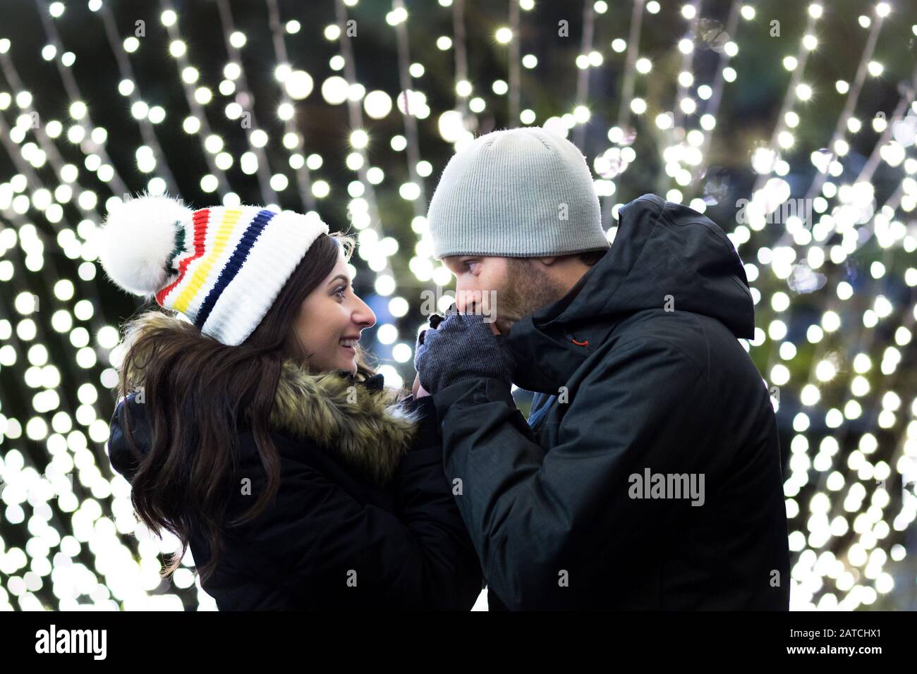 Un jeune couple caucasien attrayant en vêtements d'hiver debout devant des lumières d'arrière-plan floues embrassant et embrassant. Banque D'Images