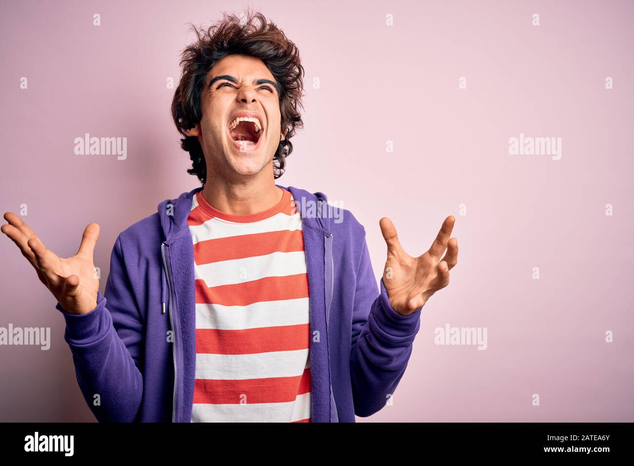 Jeune homme portant un t-shirt rayé et un sueur pourpre sur fond rose isolé folle et folle crier et hurler avec l'expression agressive et Banque D'Images