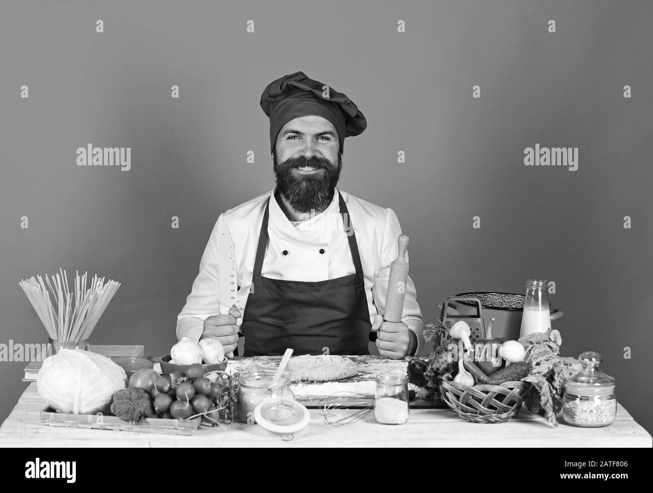Chef avec pâtes, légumes et pâte sur table. Faire cuire avec le sourire en bordeaux en uniforme maintient la goupille et le couteau à rouler. L'homme avec barbe est assis sur un comptoir sur fond rouge. Concept de cuisine maison. Banque D'Images