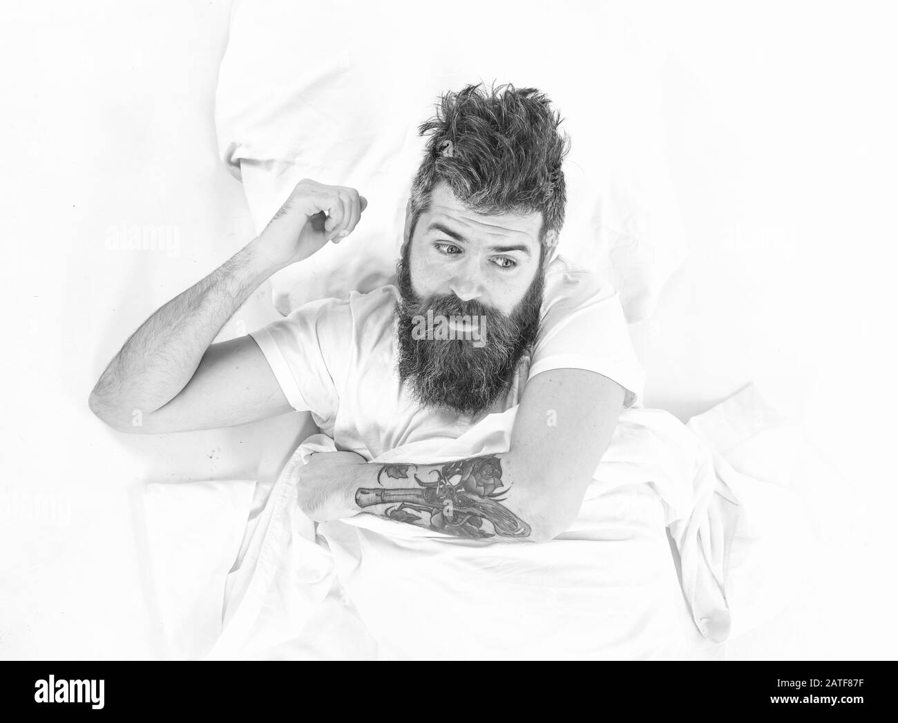 Homme avec réveil face surprise, se coucher sur lit, draps blancs, vue sur le dessus. Hipster avec barbe, moustache et cheveux bousieux se réveillant. Concept de hangover matinal. L'homme a besoin de se lever, fond blanc. Banque D'Images