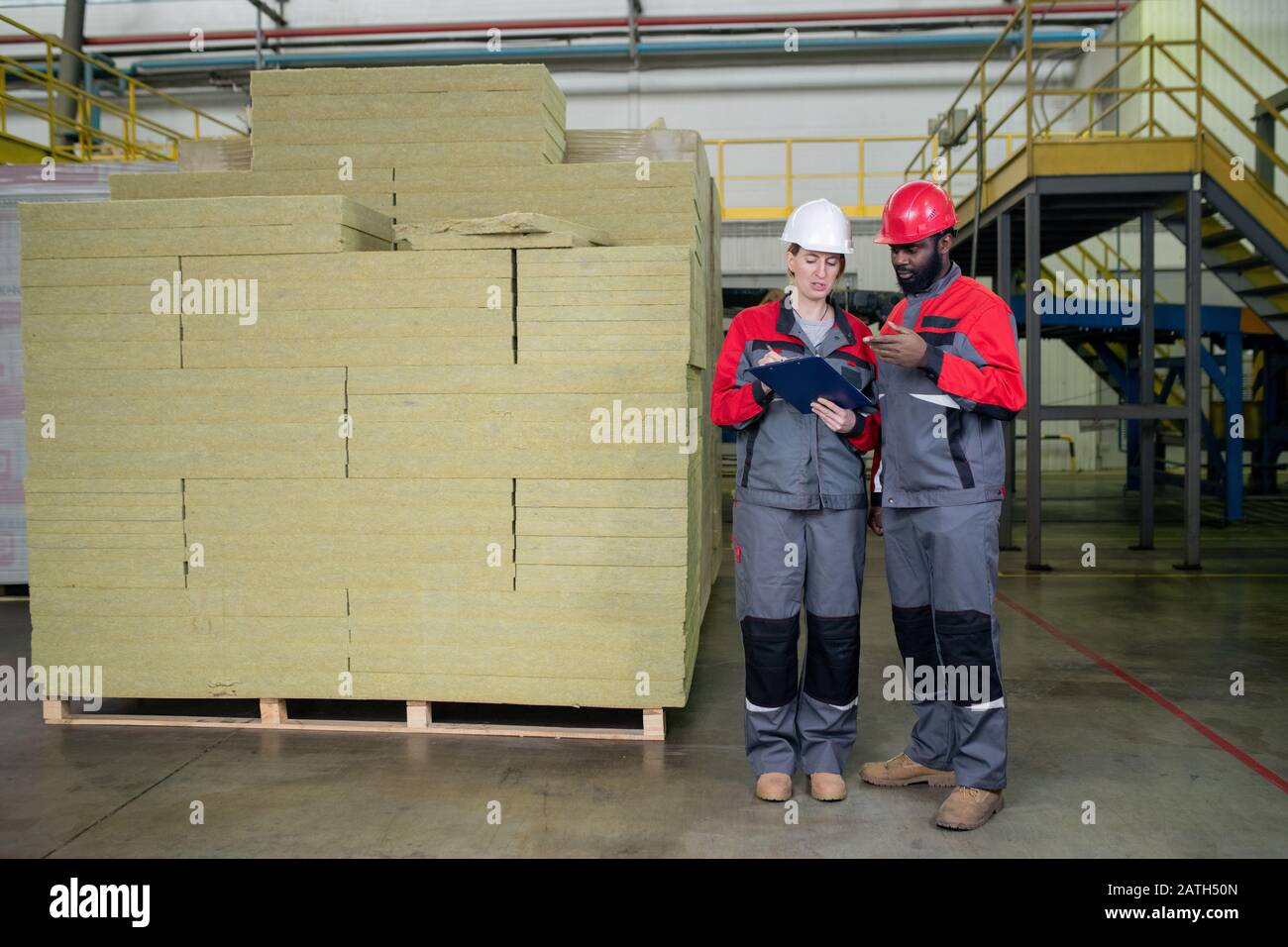 De jeunes spécialistes de la construction multiethnique dans les casques se tenant à la pile de laine de pierre et discutant des données en entrepôt Banque D'Images
