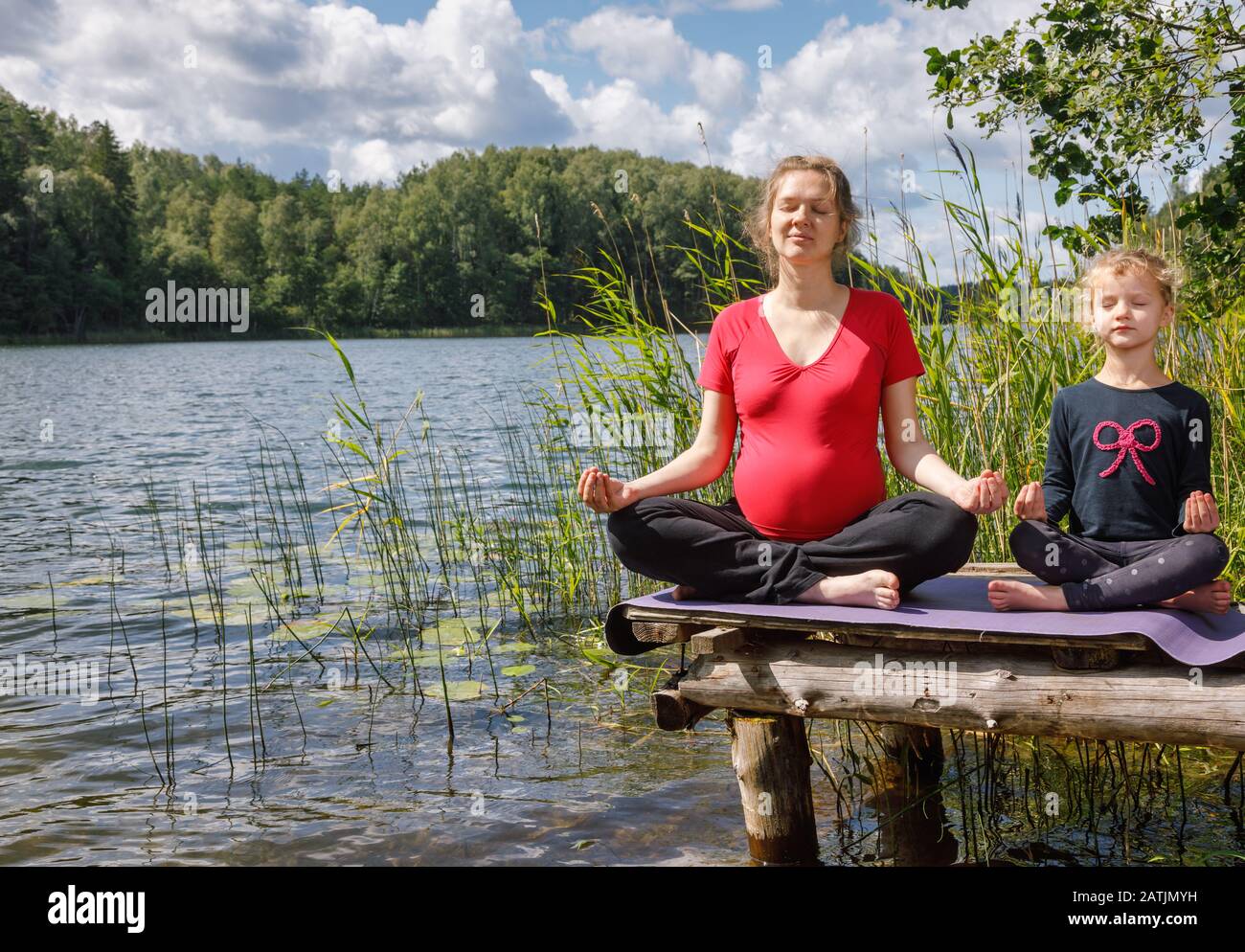 Jeune femme enceinte portant une robe sportive faisant de l'exercice de yoga assis en position lotus sur une jetée en bois à un lac forestier méditant. Petite fille Banque D'Images