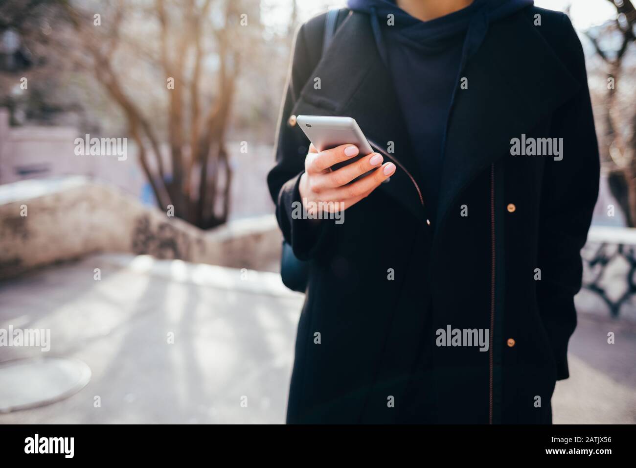 Jeune femme portant un élégant manteau noir à l'aide d'un smartphone dans la rue de la ville le jour ensoleillé du printemps, gros plan. Banque D'Images