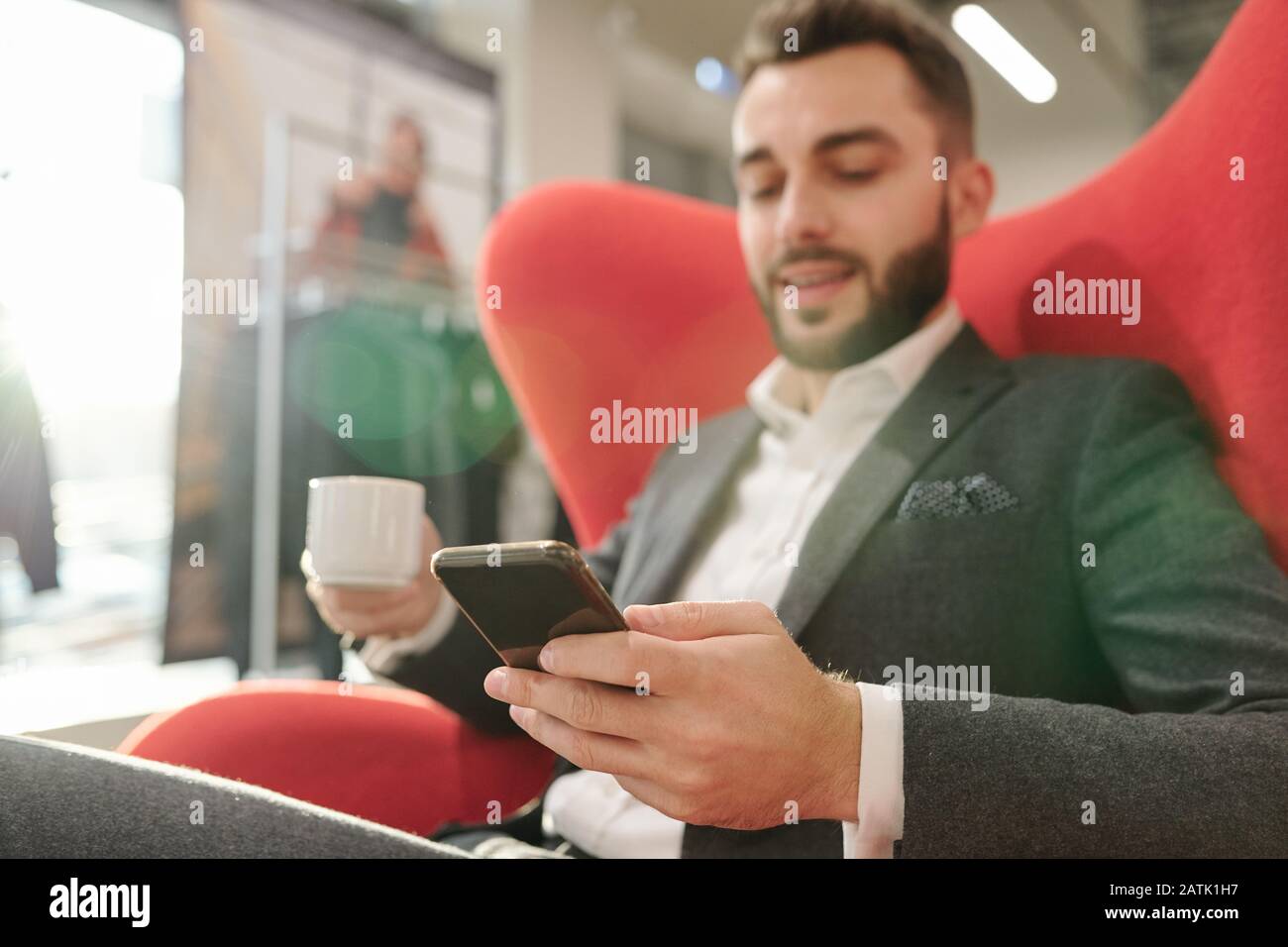 Content jeune homme d'affaires dans une élégante veste assise dans un fauteuil confortable et boire du café tout en surfant sur le téléphone dans la boutique de vêtements Banque D'Images