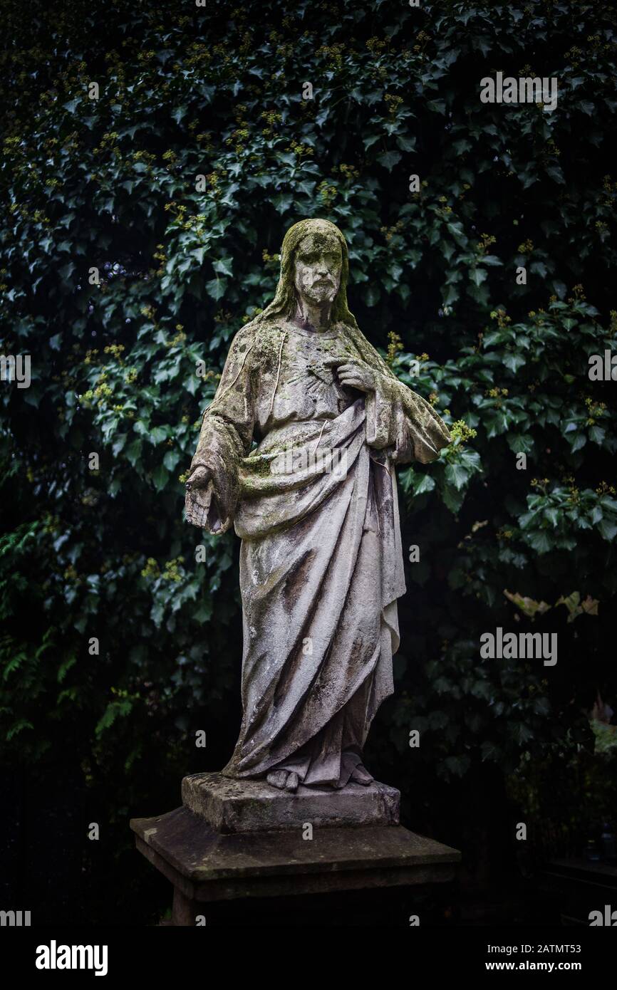 Jésus Christ sculpture en pierre contre les plantes vert foncé fond sombre dans la nécropole ancienne, cimetière de Rakowicki, Cracovie, Pologne Banque D'Images